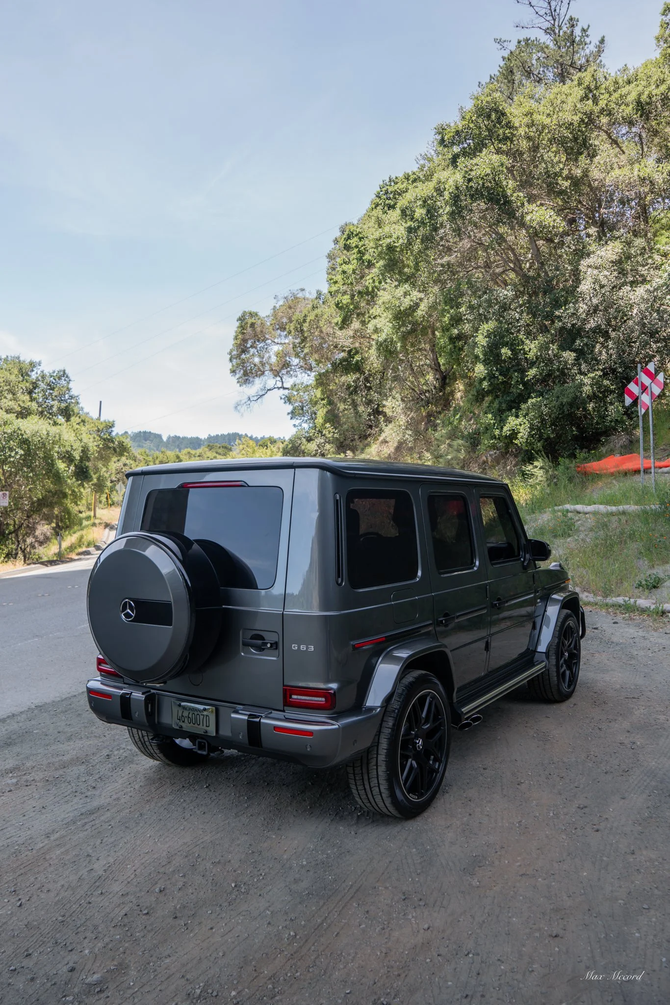 A black Mercedes-Benz G63 SUV parked on the side of a rural road with trees, bushes, and construction signs in the background.