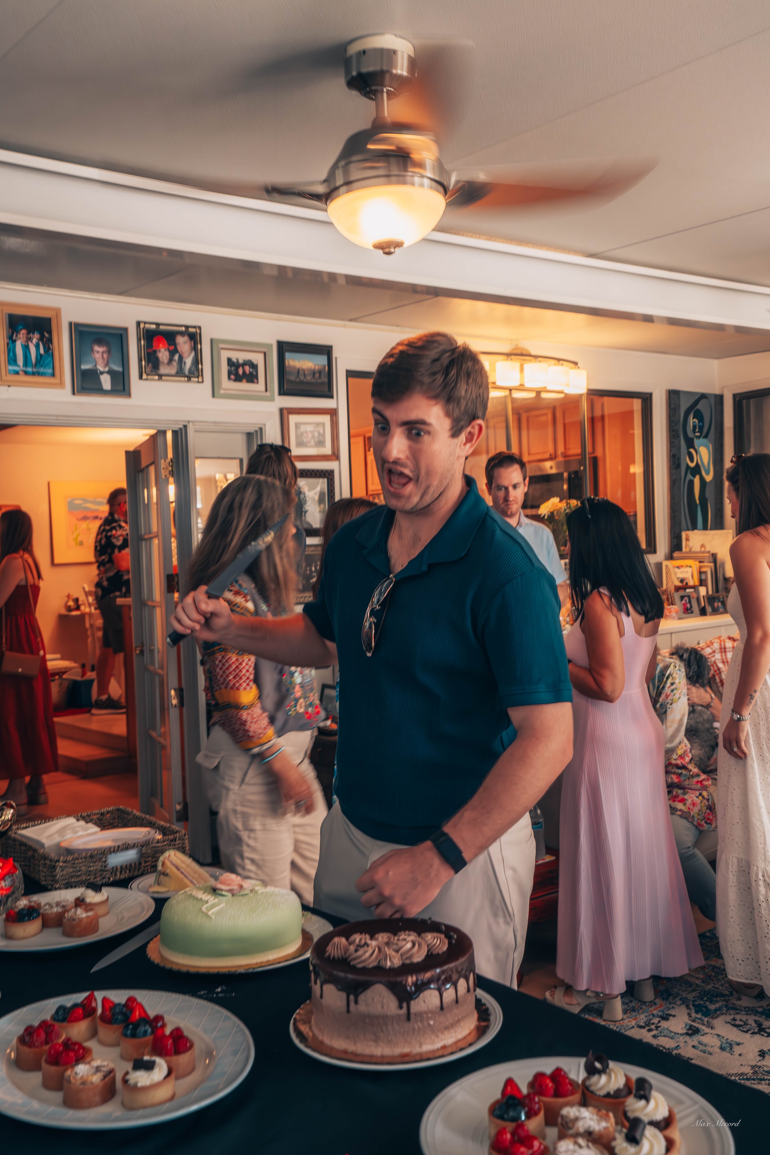 A man at a birthday party is holding a knife and standing in front of a table with various cakes and desserts. The party is in a decorated living room with multiple people in the background.