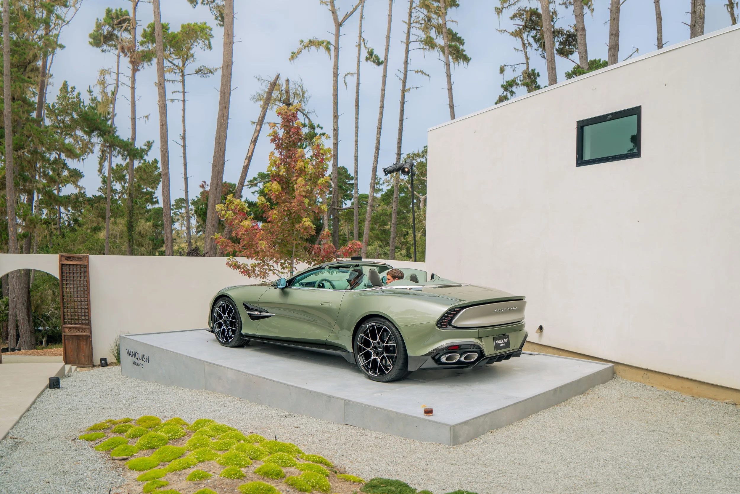 A green Aston Martin V12 Speedster displayed on a platform outdoors with trees in the background.
