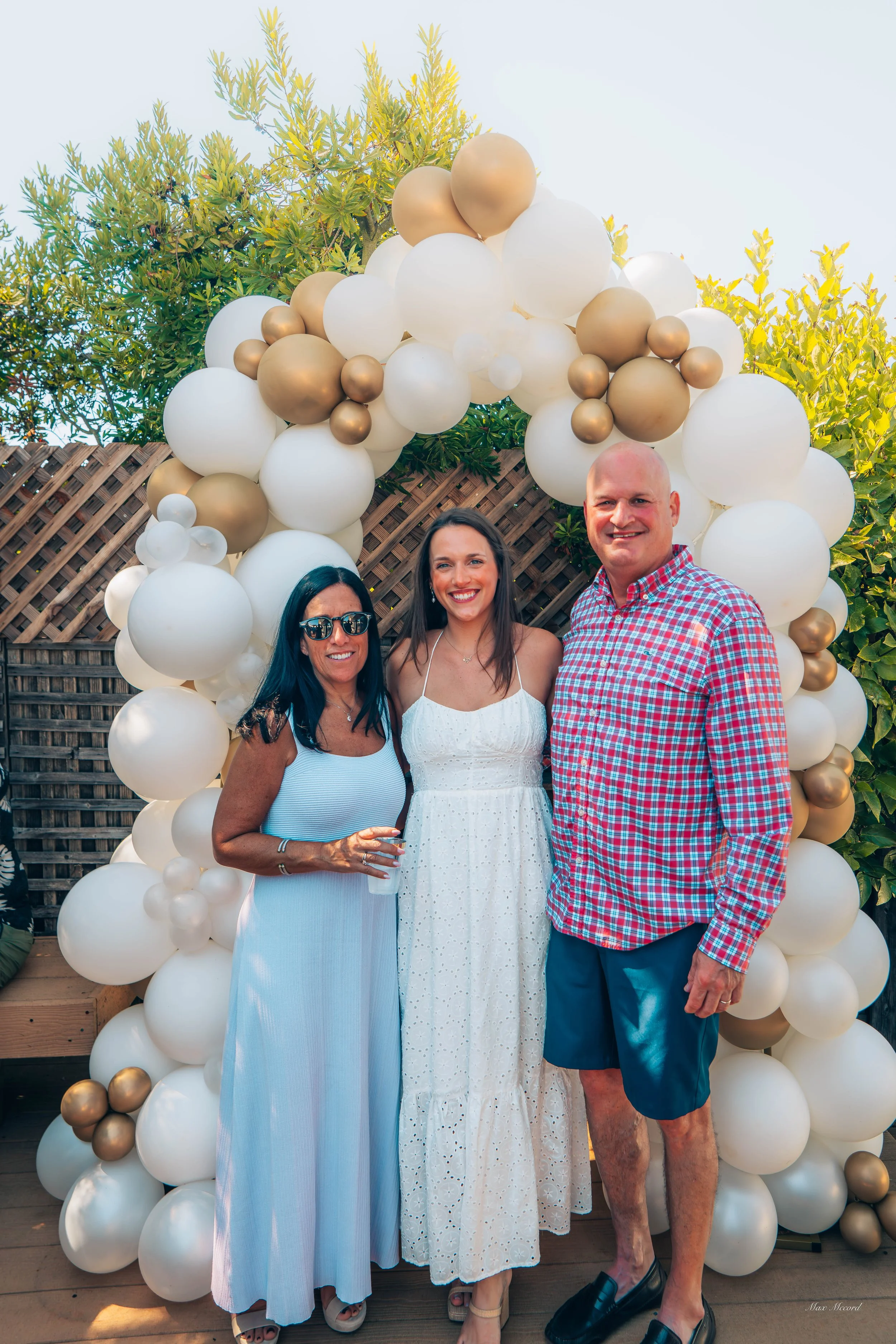 Three people standing in front of a balloon arch outdoors. The arch is decorated with white and gold balloons. The group includes an older woman wearing sunglasses and a light blue dress, a young woman in a white sundress, and a man in a red and blue