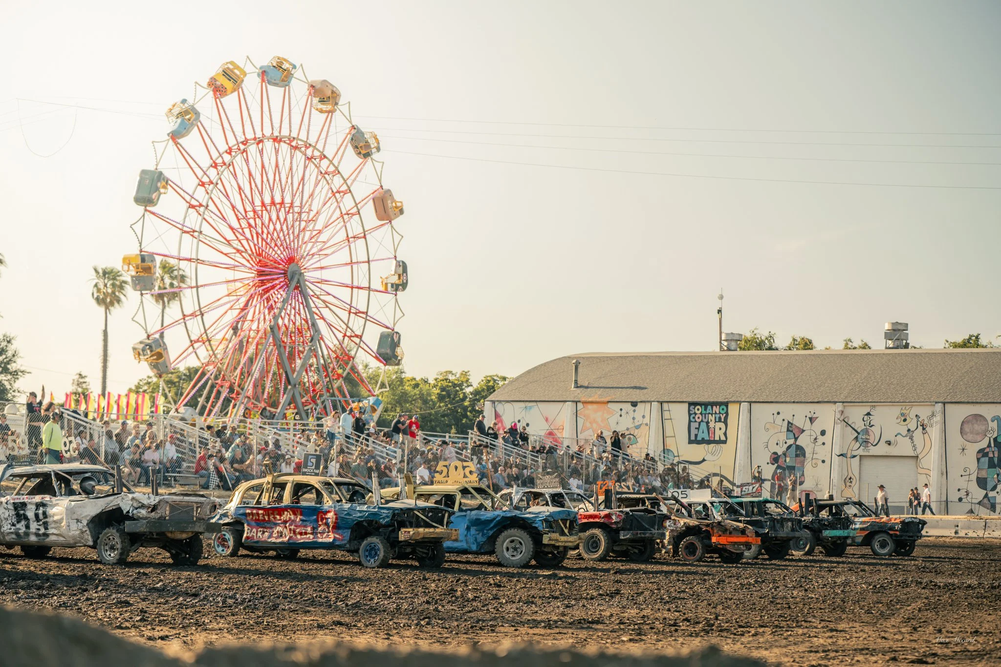 A carnival with a large colorful Ferris wheel, a building with 'Solano County Fair' signage, and rows of wrecked cars in the foreground.