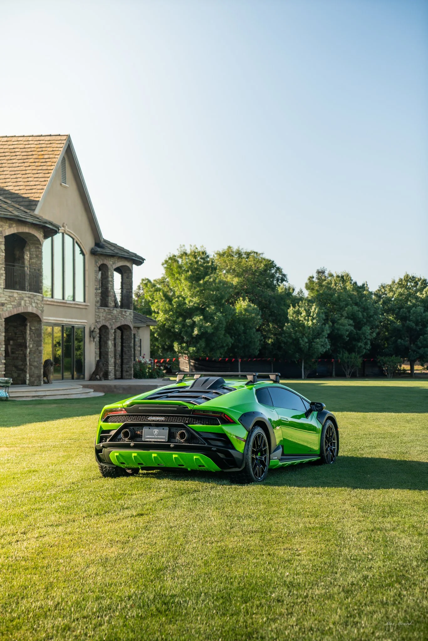 A bright green sports car parked on a manicured lawn outside a large house with stone and stucco exterior and large windows, with trees in the background.