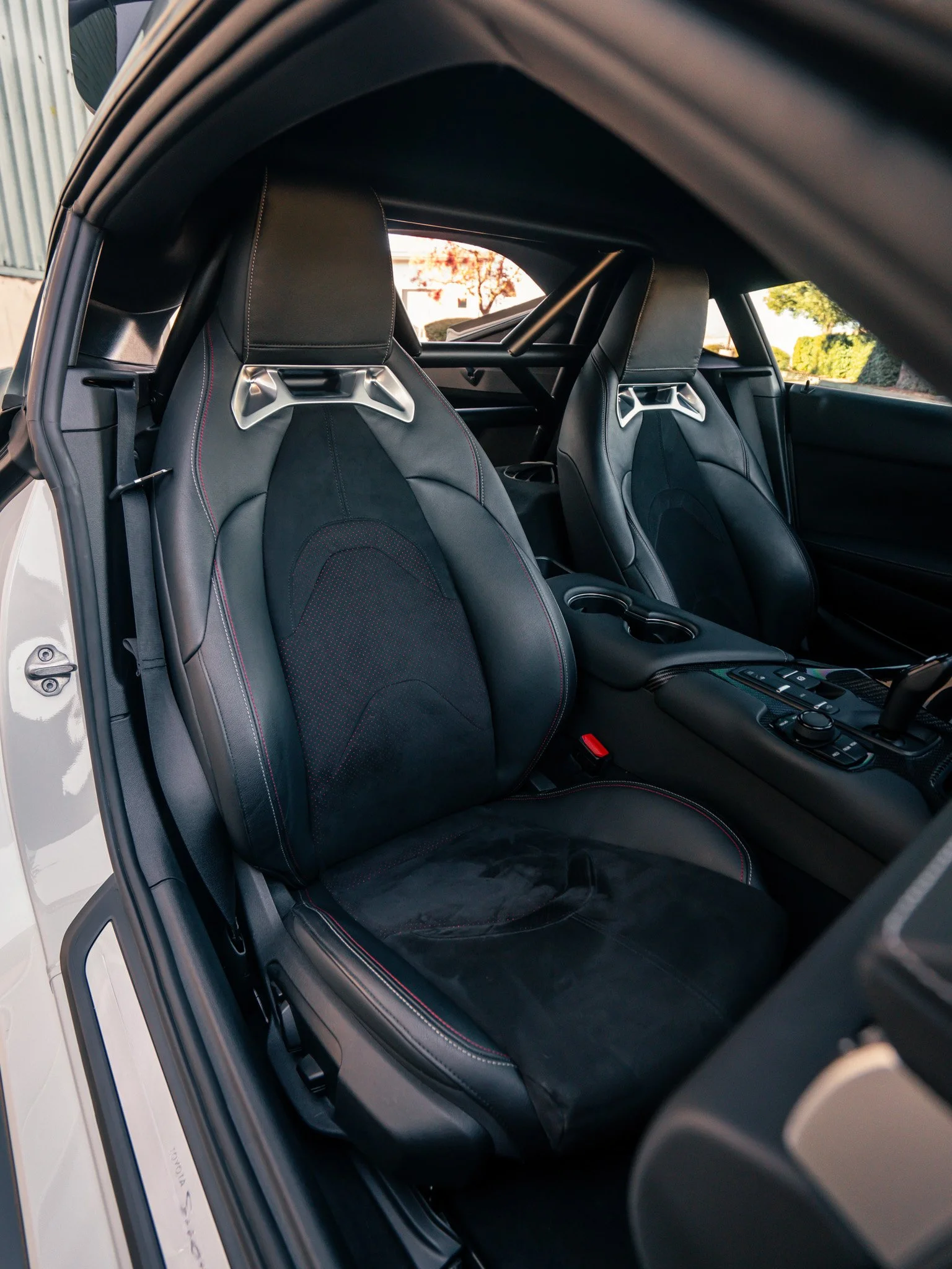 Inside a car, two sports seats with black leather upholstery, red stitching, and metal racing harness attachments. The interior appears modern with a focus on performance.