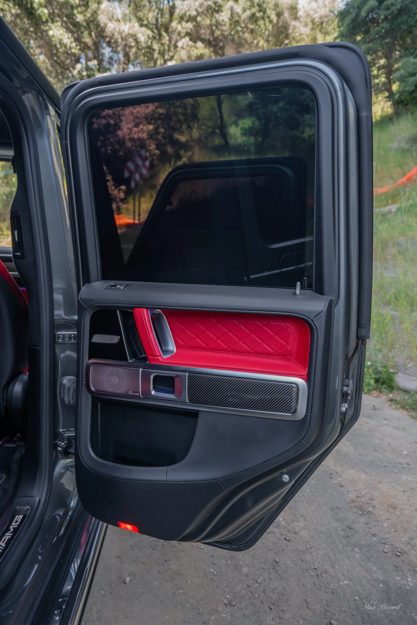 The open door of a black luxury vehicle, showing red, black, and carbon fiber interior door panel with speakers and controls in an outdoor setting.