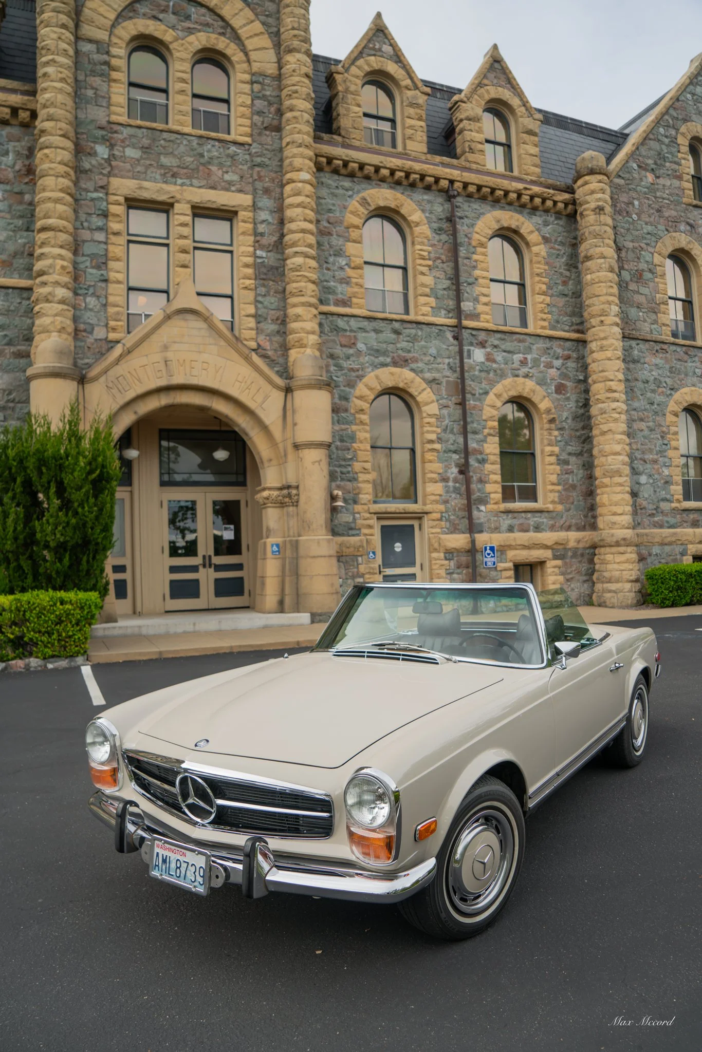 A vintage cream-colored Mercedes-Benz convertible parked outside Montgomery Hall, a historic stone building with arched windows and an ornate stone entrance, with some greenery nearby.