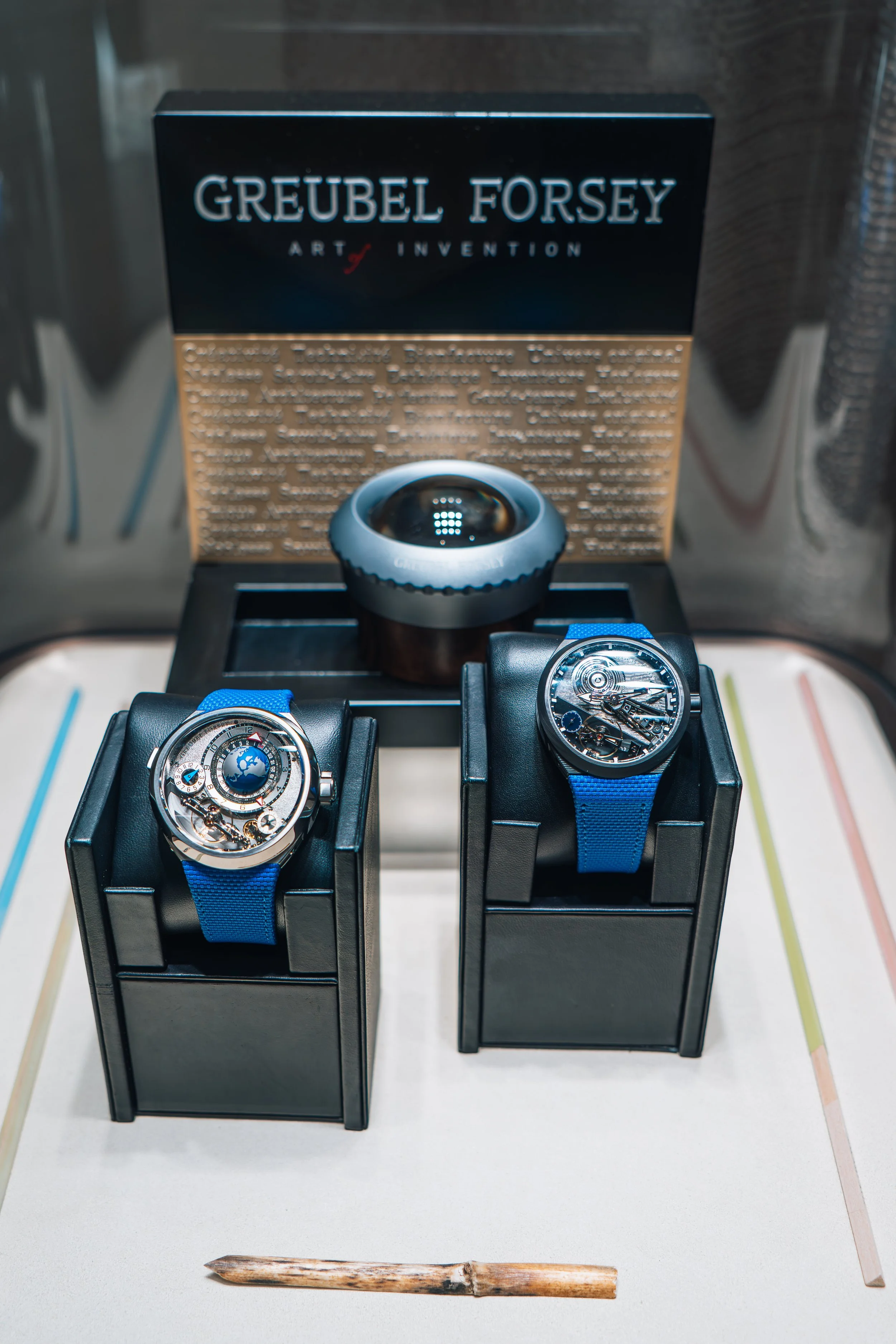 Two luxury watches with blue straps displayed on black stands, a camera with a reflective lens in the background, and a box with the text 'Greubel Forsey' and information about the brand behind the watches.