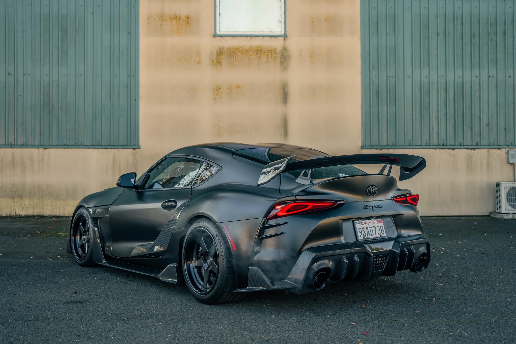 A matte black Toyota Supra sports car with a large rear spoiler and custom racing wheels, parked on an asphalt surface against a weathered building backdrop.