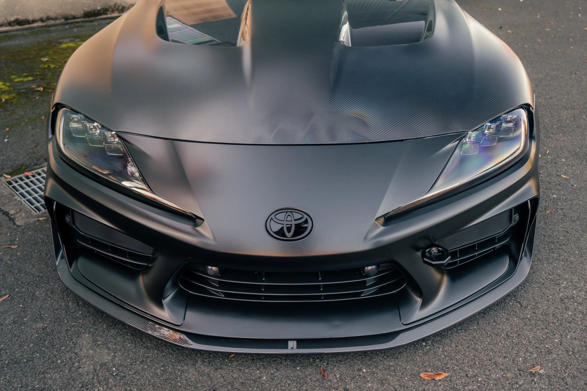 Front view of a matte gray Toyota Supra sports car with sleek headlights and a prominent hood scoop, parked on asphalt with some fallen leaves.