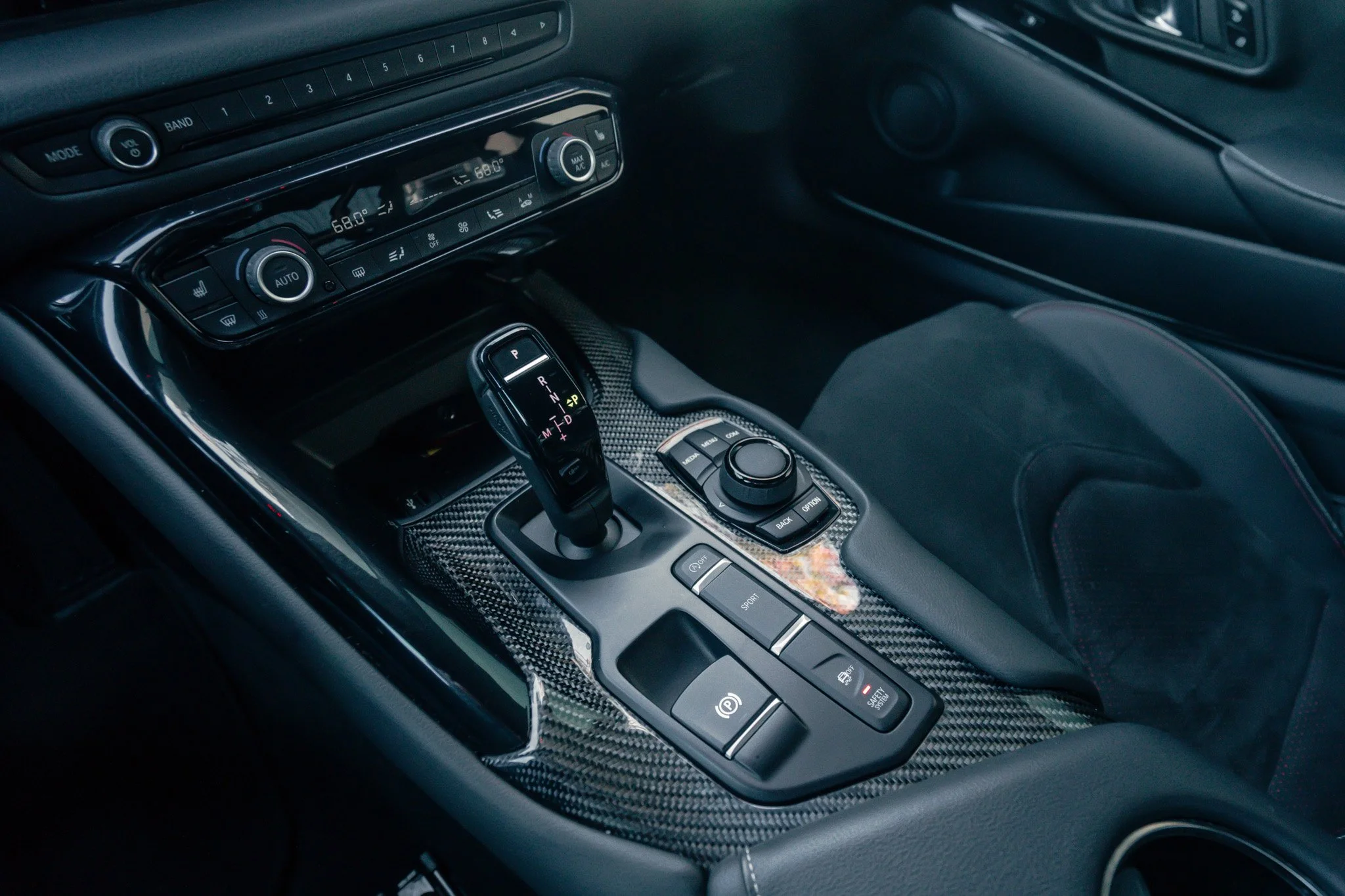 Interior of a car showing a center console with a gear shift, control knob, electronic parking brake, and various buttons, with a carbon fiber finish.