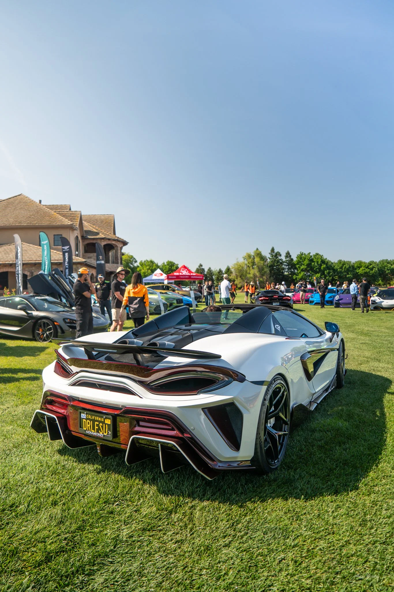 A white sports car with a custom spoiler parked on a grassy field during a car event, with other sports cars, attendees, and tents in the background under a clear blue sky.