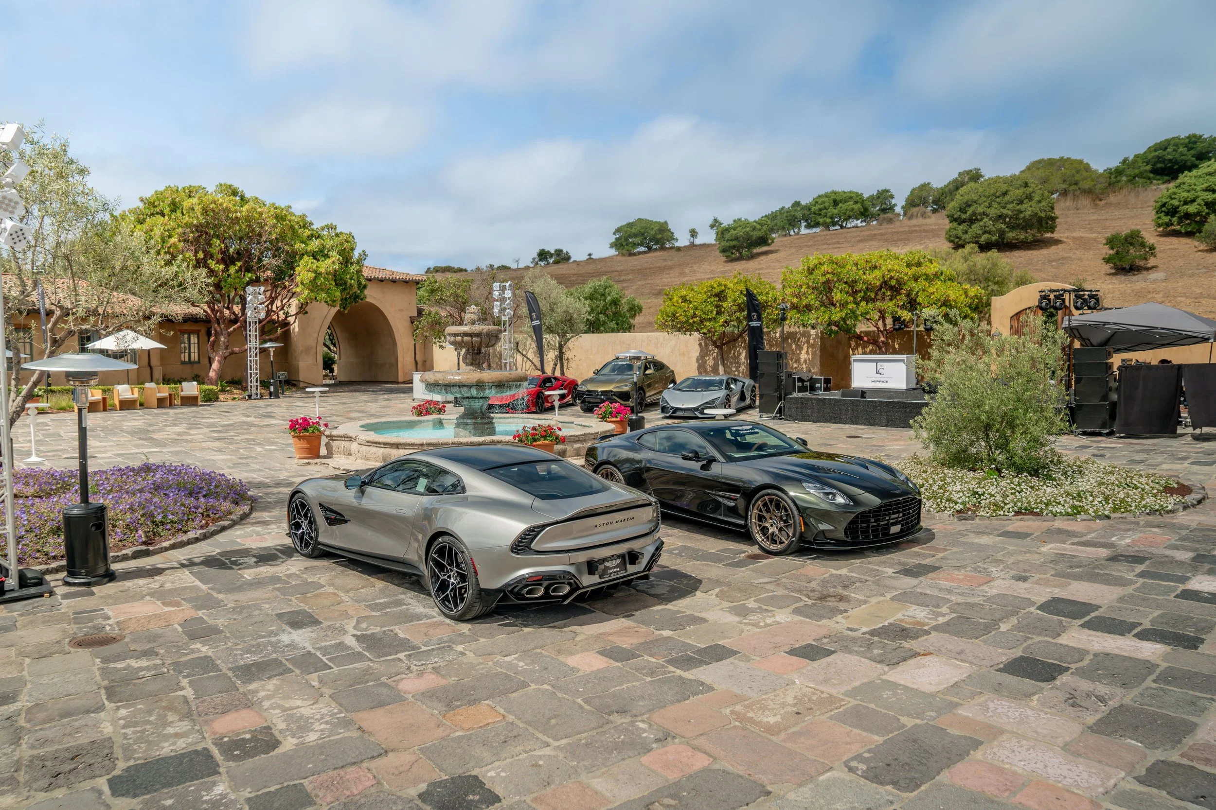 Luxury cars parked in an outdoor courtyard with a fountain, trees, and a stage setup, possibly for an event, on a sunny day with a cloudy sky.