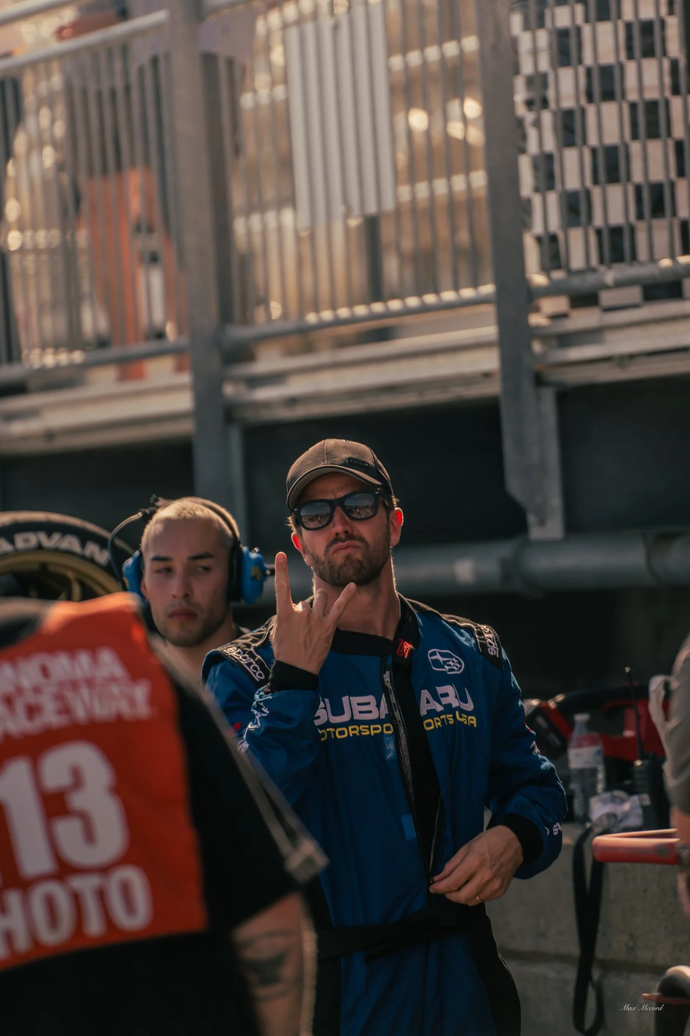 A man in racing gear, wearing sunglasses and a cap, making a rock on hand gesture at a racing event.