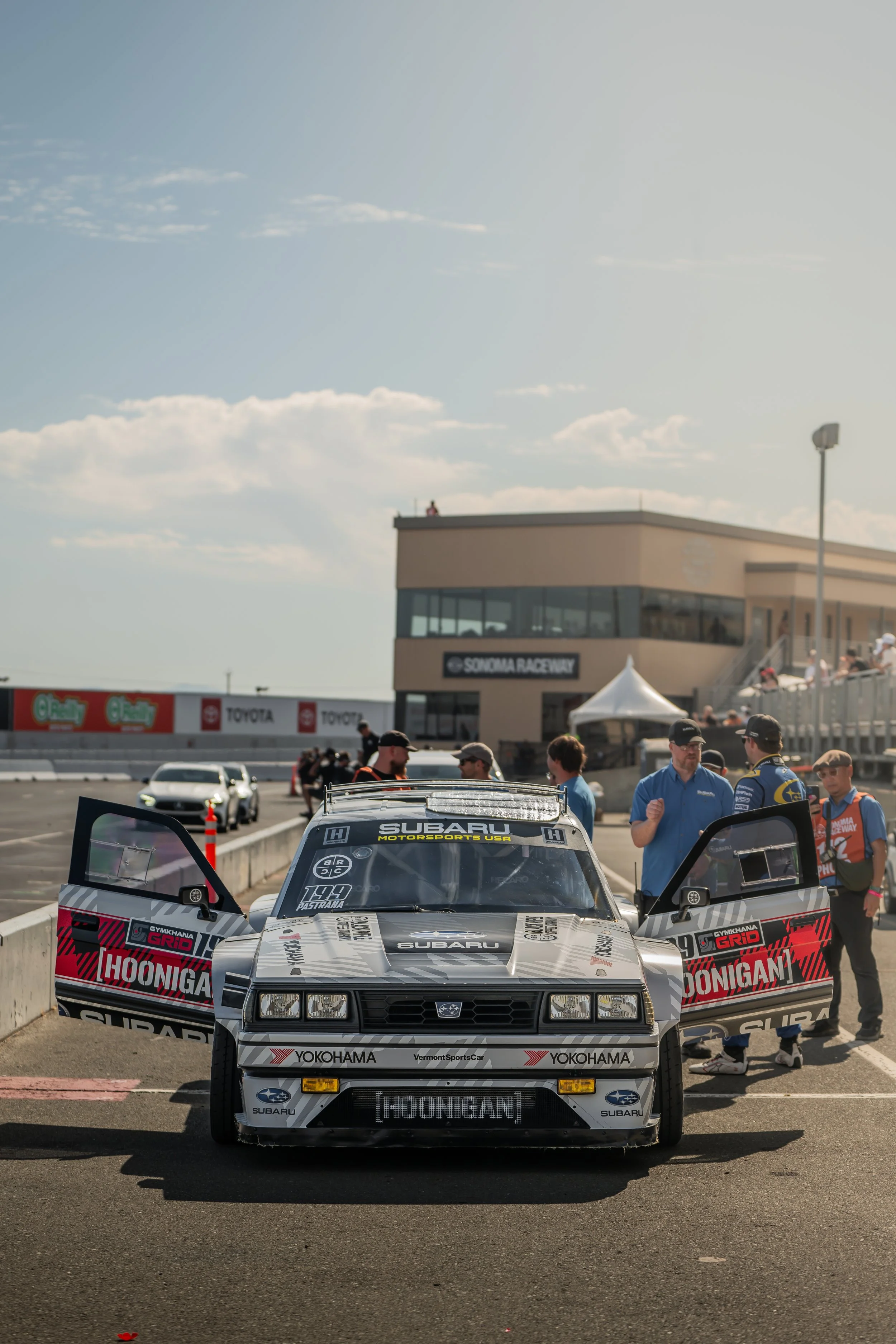 A race car in the pit lane at Sonoma Raceway with team members and officials standing nearby on a sunny day.