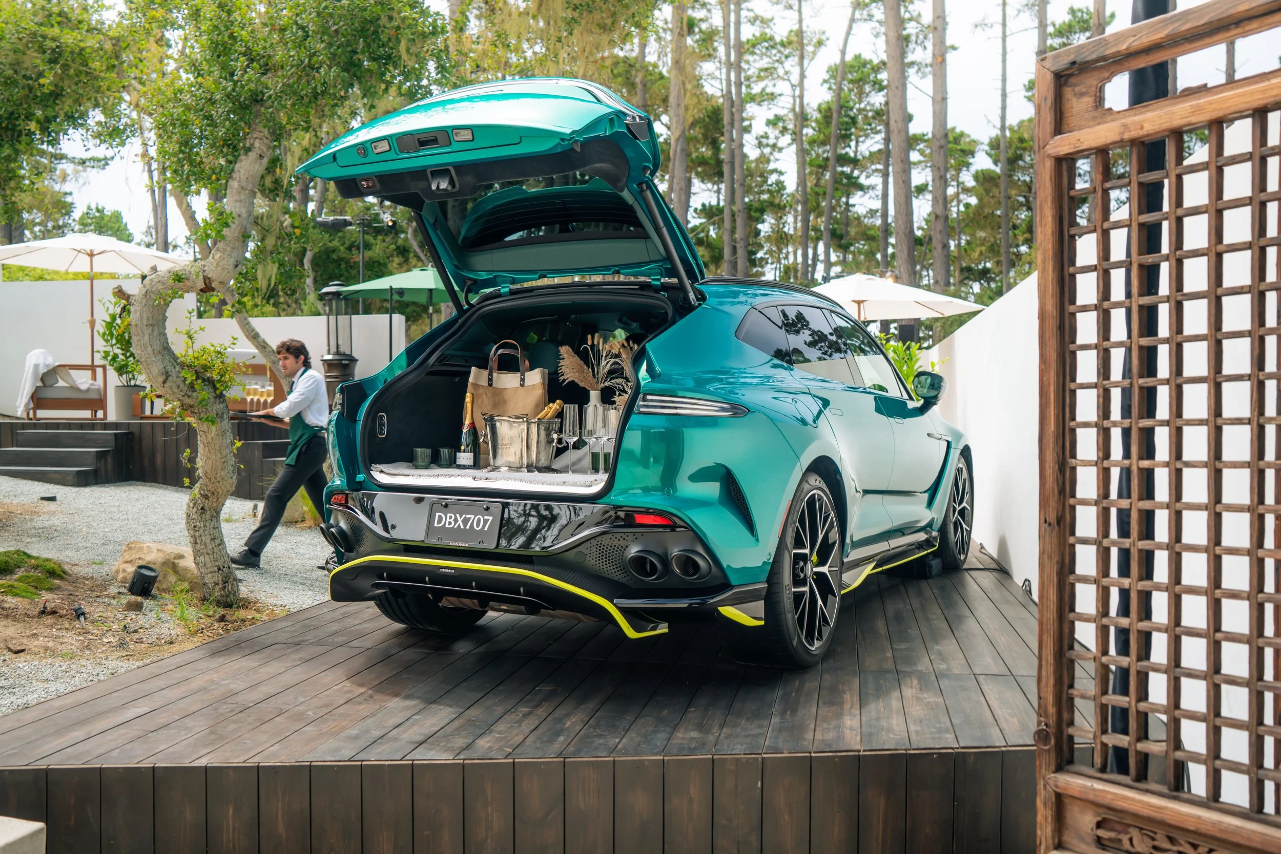 An electric green luxury car with its trunk open, parked on a wooden platform outdoors, with a man in the background near a seating area and trees.