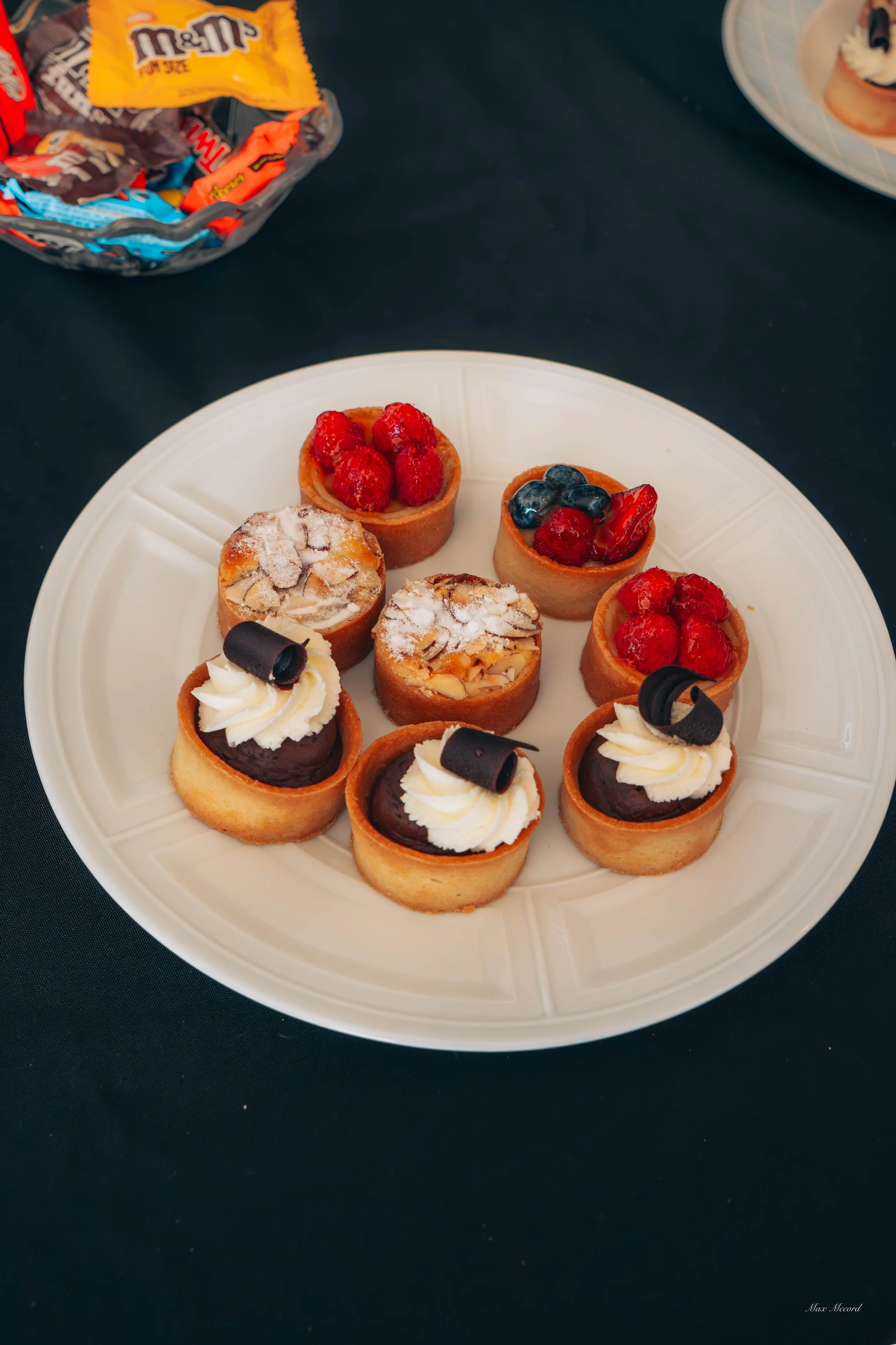 A white plate with eight assorted mini tarts topped with berries, chocolate, and whipped cream, on a black table with a bag of candy and a dessert plate partially visible in the background.