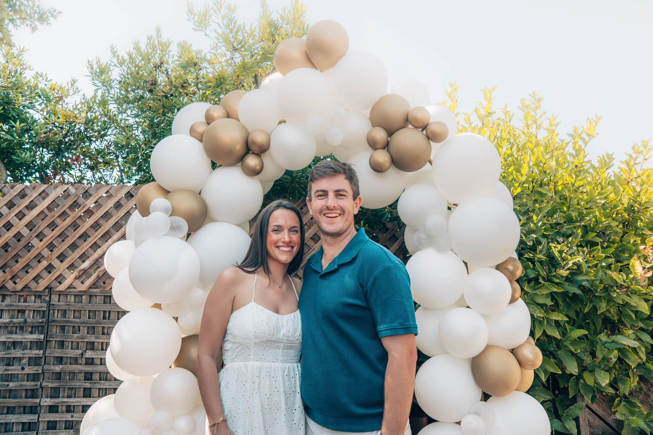 A young couple standing in front of a decorative balloon arch made of white and gold balloons, outdoors with green foliage and a wooden fence in the background.