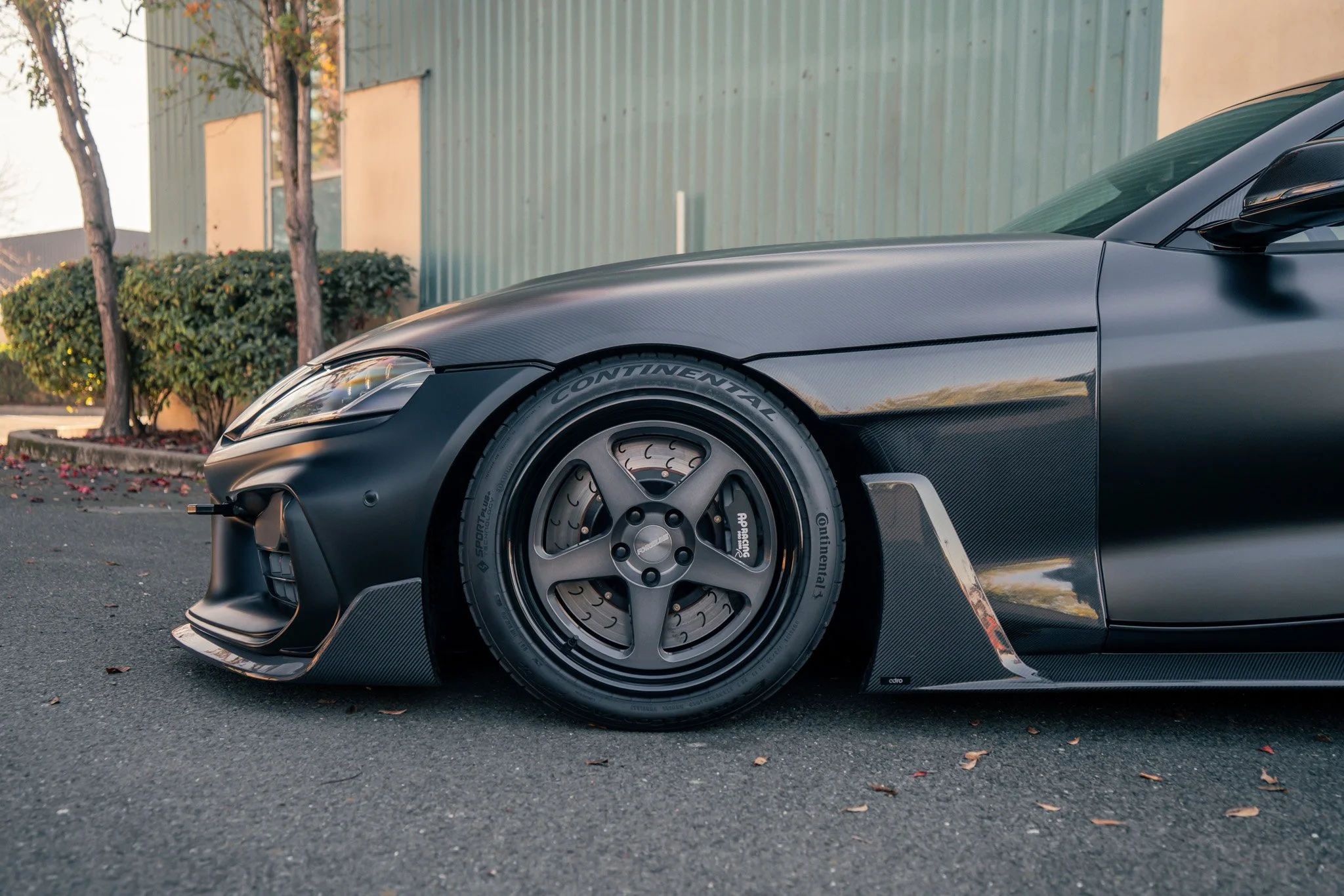 Close-up of a sleek, black sports car with a carbon fiber front splitter, racing slick tires, and a low aerodynamic profile parked on a street with trees and a building in the background.