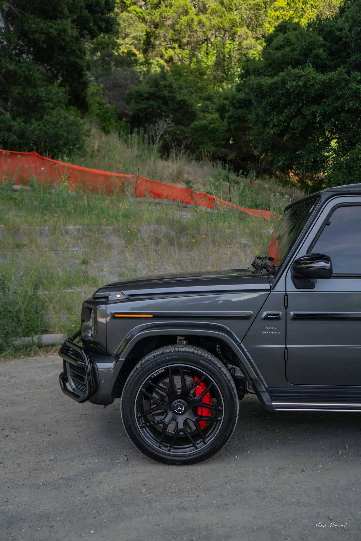 Close-up of the front part of a black Mercedes-Benz G-Class SUV with red brake calipers, parked on a dirt road with greenery and trees in the background.