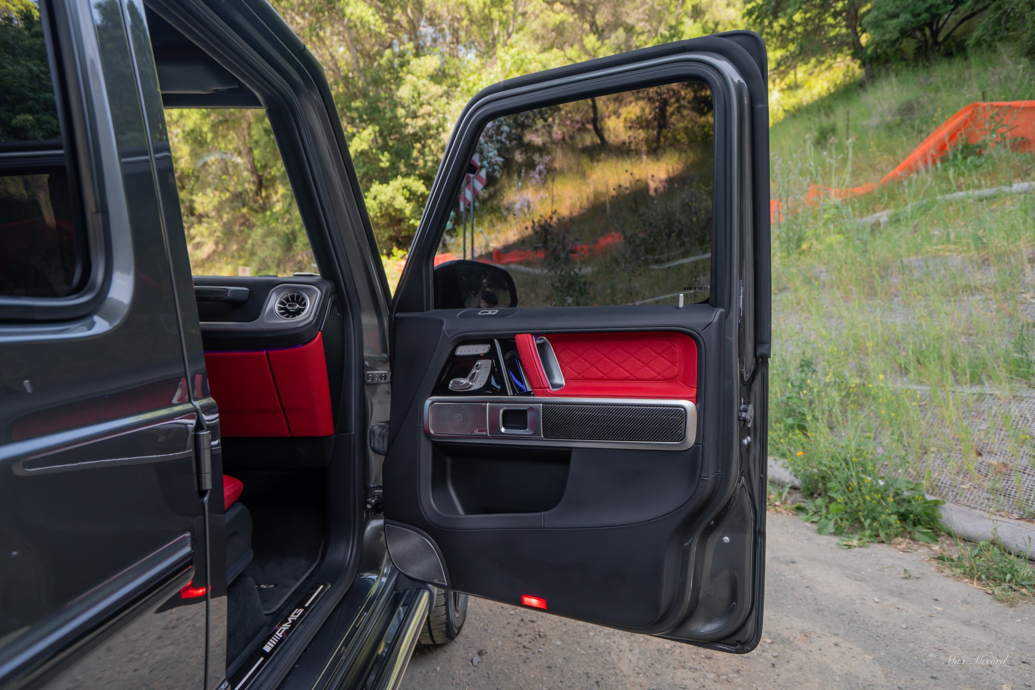Open car door revealing black interior with red accents, set outdoors with greenery and orange barrier in the background.