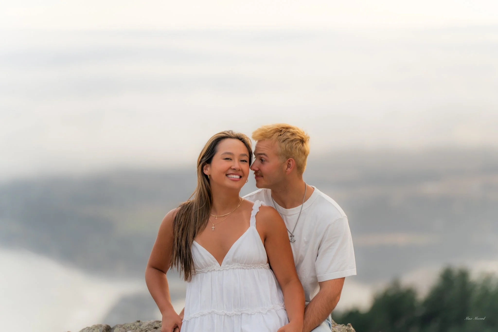 Young woman in a white dress and young man in a white t-shirt close together outdoors, with a blurred natural background.