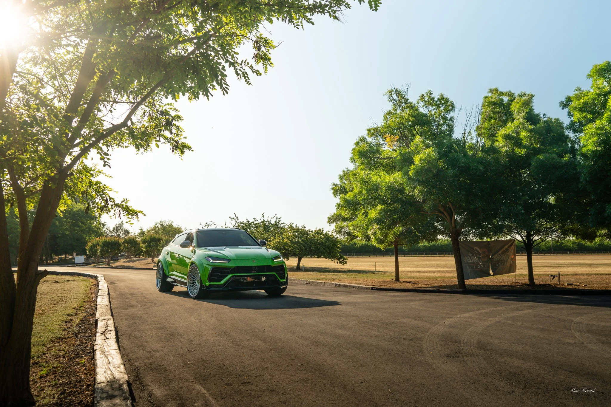 A green luxury SUV parked on a curved asphalt driveway in a park-like setting with trees and open grassy fields on a sunny day.
