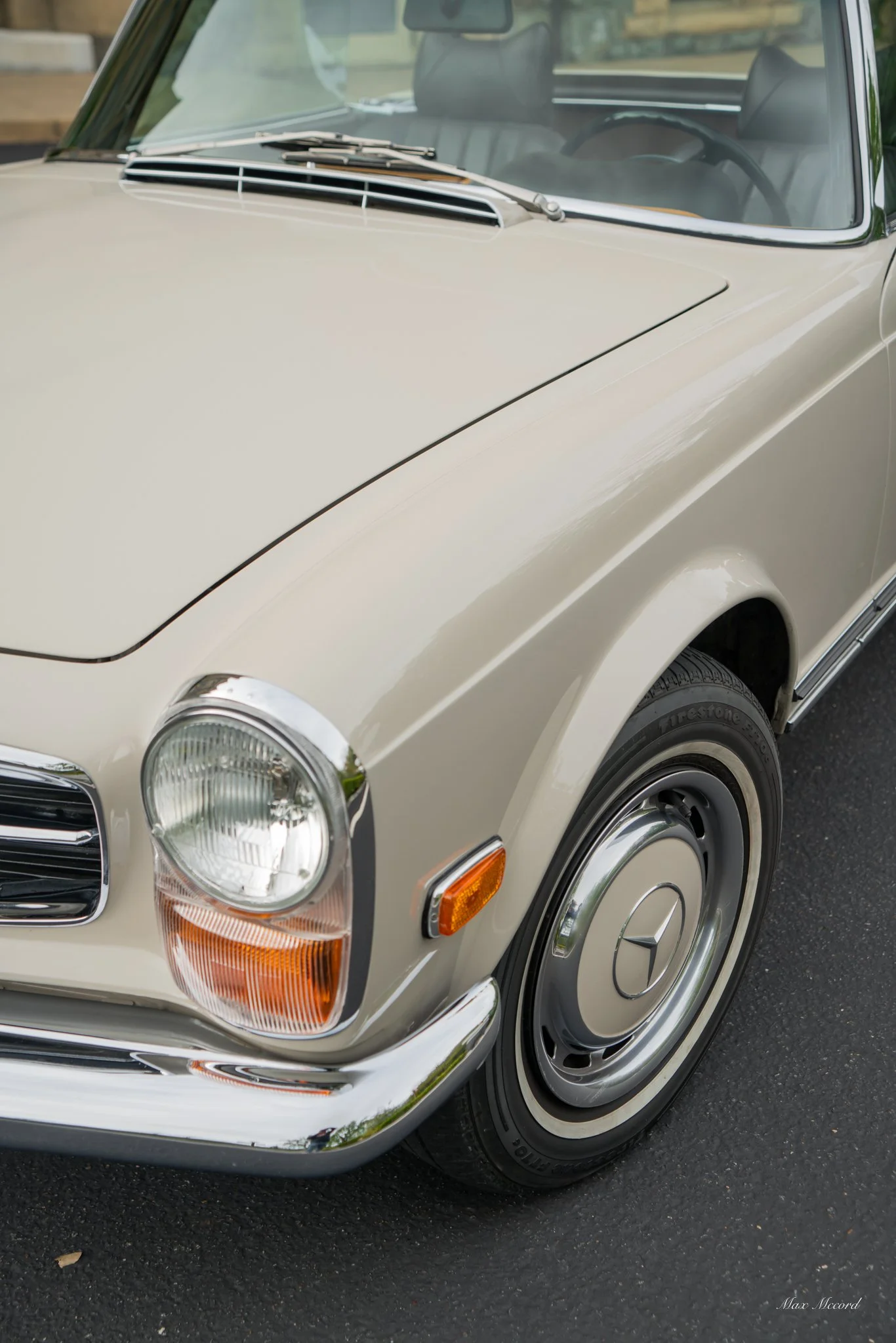 Close-up of a vintage beige Mercedes-Benz car showing the front left headlight, bumper, wheel, and part of the windshield.