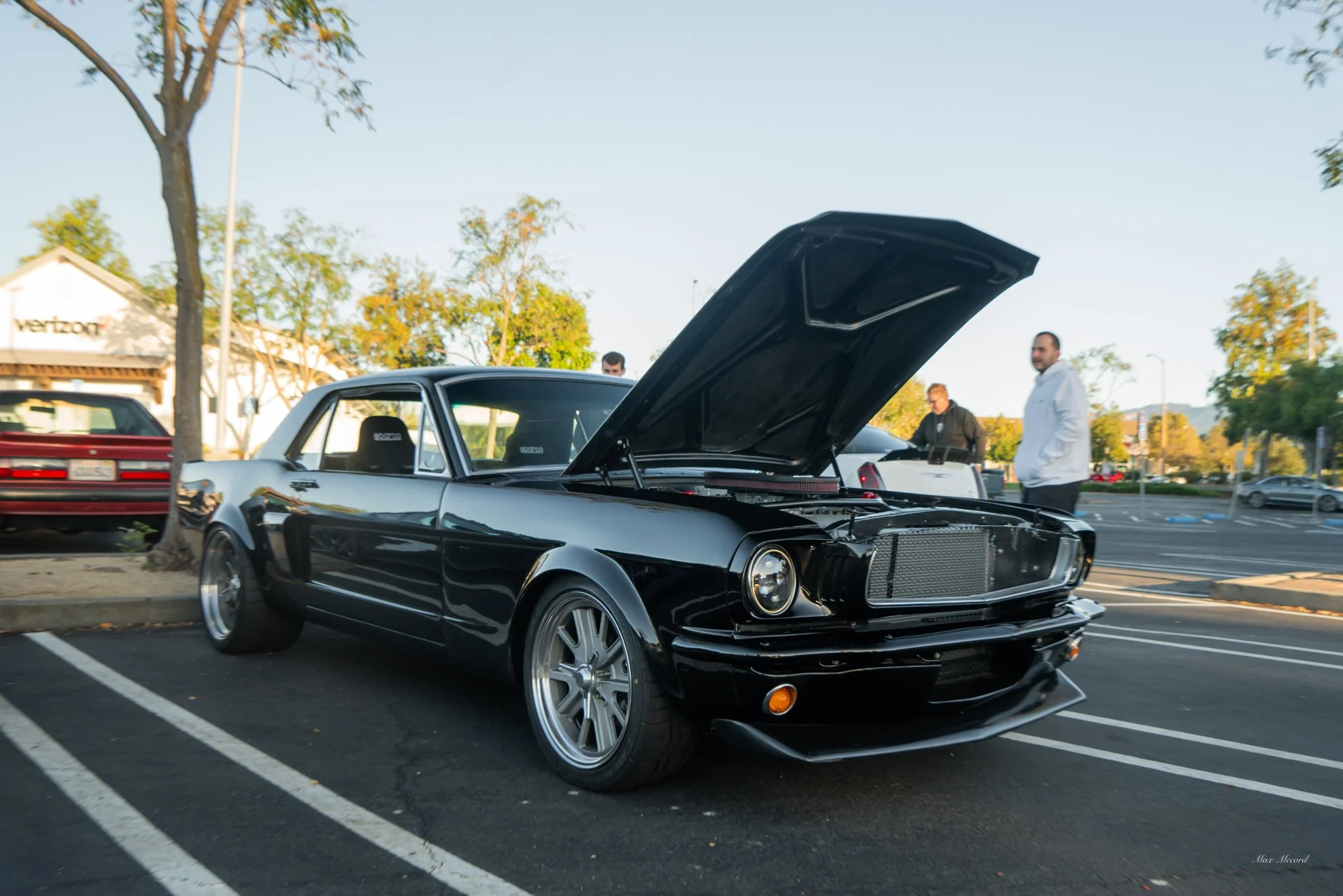 Black classic car with hood open at a parking lot, with three people standing nearby and trees in the background.