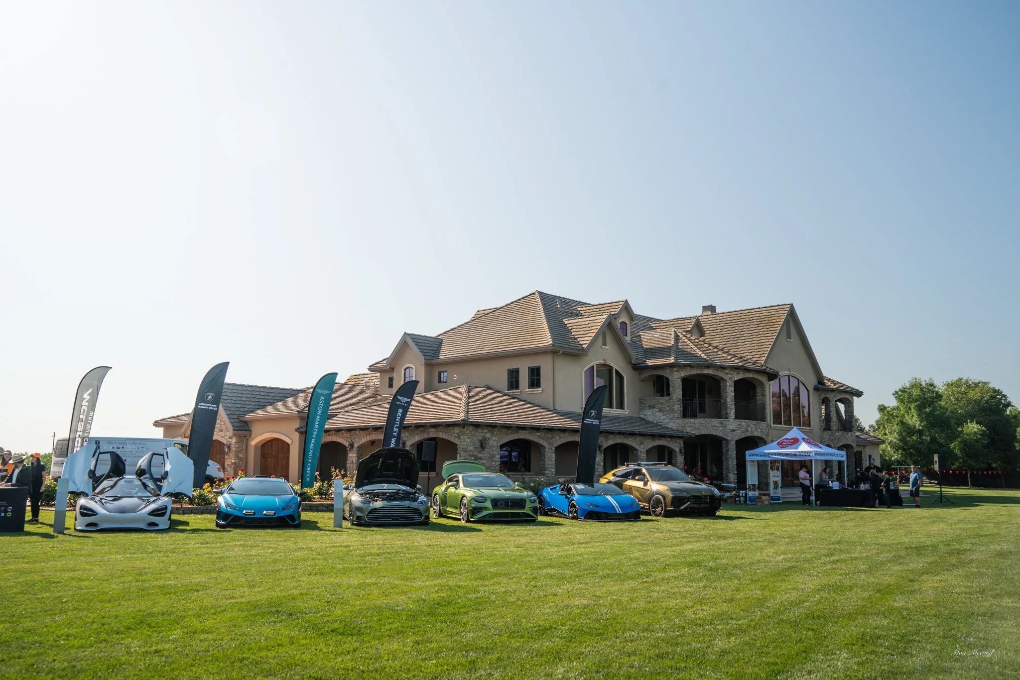 Luxury cars displayed on a lawn outside a large house with stone and stucco exterior, with promotional flags and a tent at a car event on a sunny day.