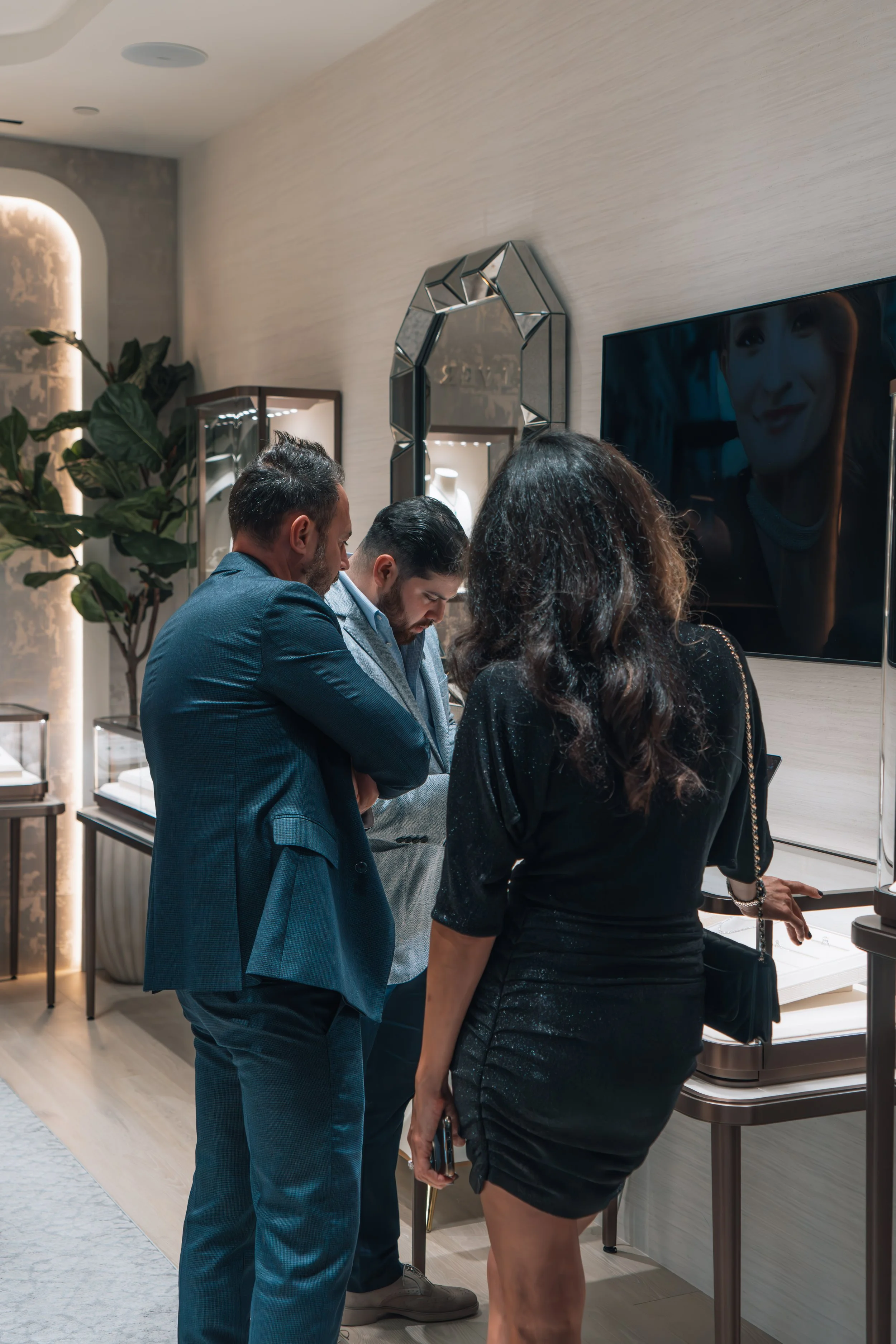 Three people looking at jewelry in a store, with display cases and a large mirror behind them.