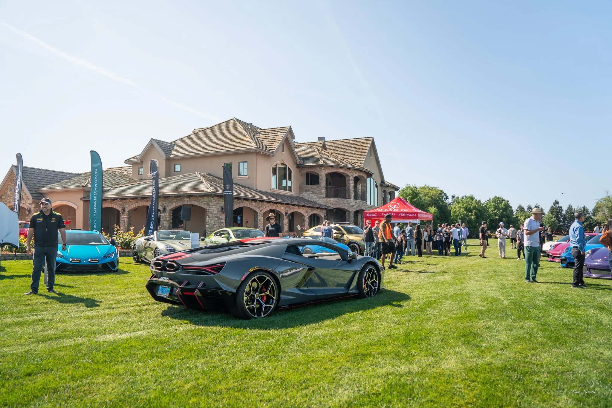 A luxury car show on a manicured lawn in front of a large house, featuring a sleek black Lamborghini supercar, several colorful sports cars, and people walking around and viewing the vehicles under a clear blue sky.