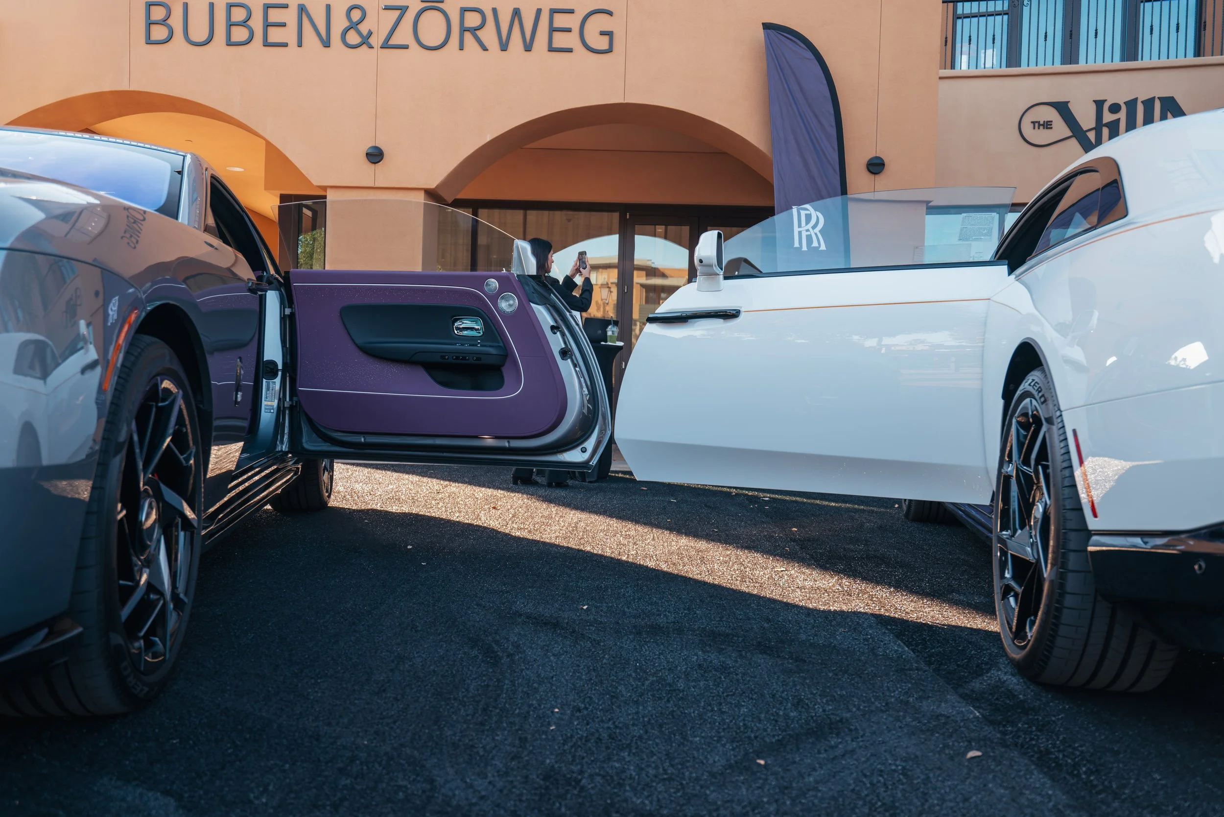 Two luxury cars, a dark blue/gray and a white one, parked outside a building with the sign "BUBEN & ZORWESG". The purple door of the dark blue car is open, and a woman is standing nearby taking a photo.