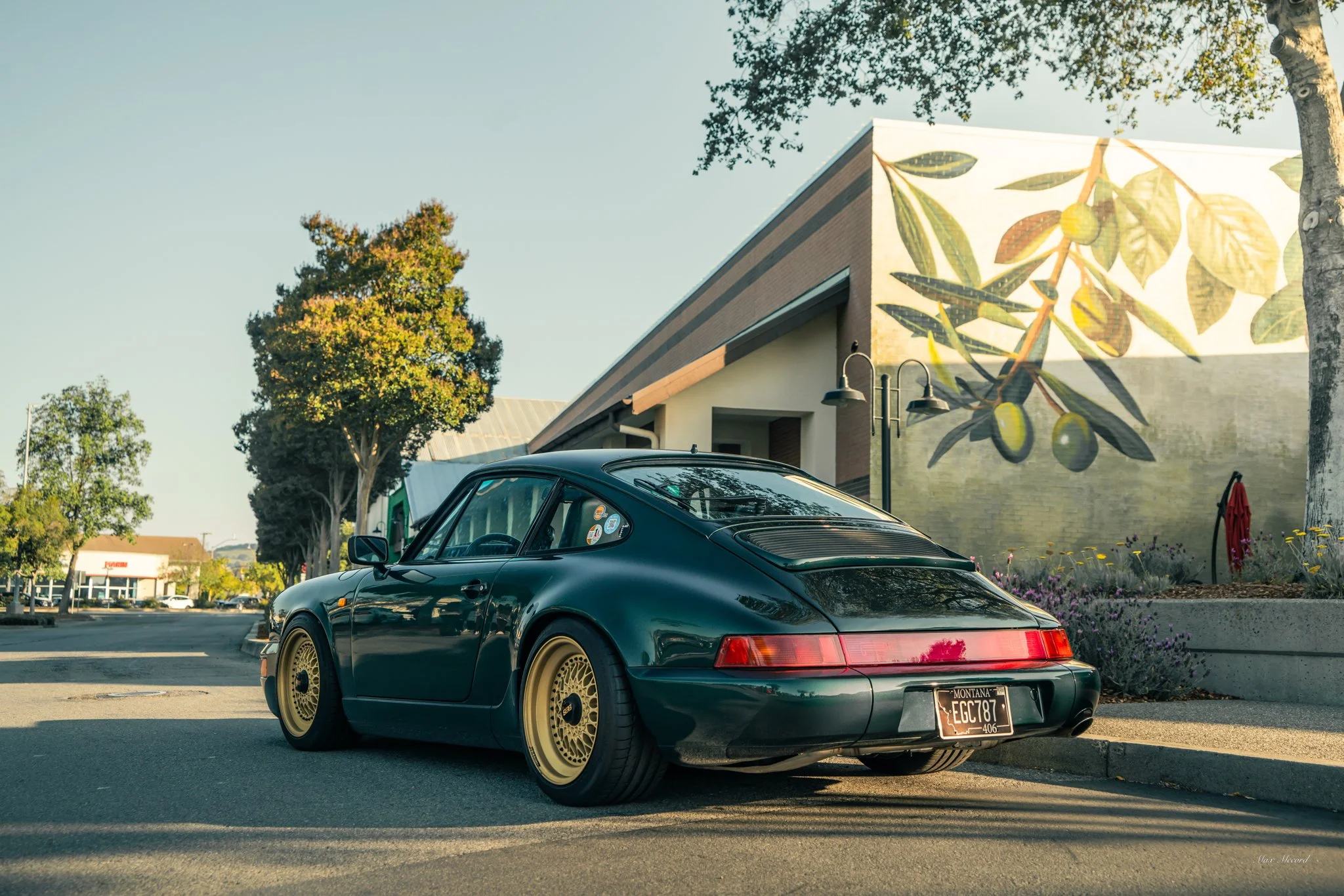 Black vintage Porsche with gold wheels parked on a street with a large building featuring a mural of green olives painted on its wall.