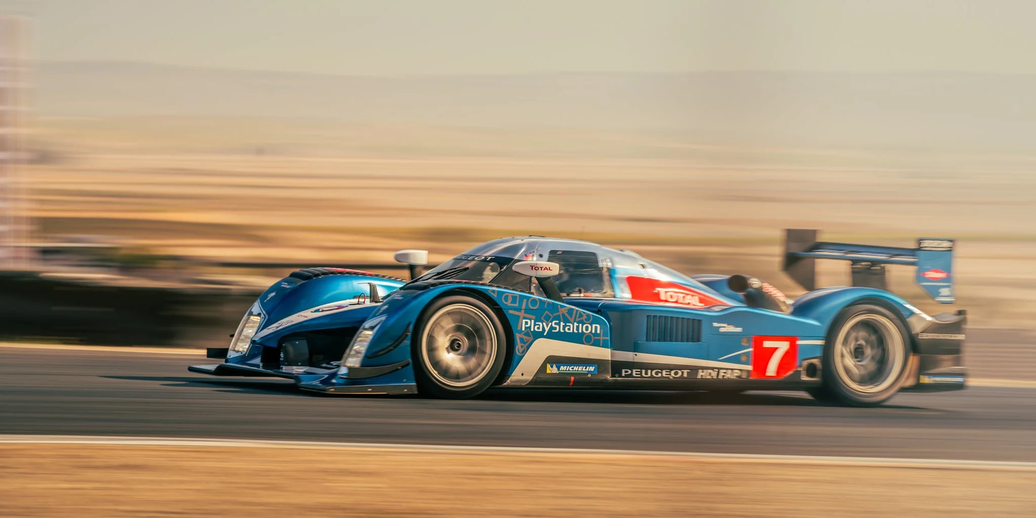 A blue Peugeot race car with PlayStation and Total logos, number 7, speeding on a racetrack during daytime, with motion blur background.