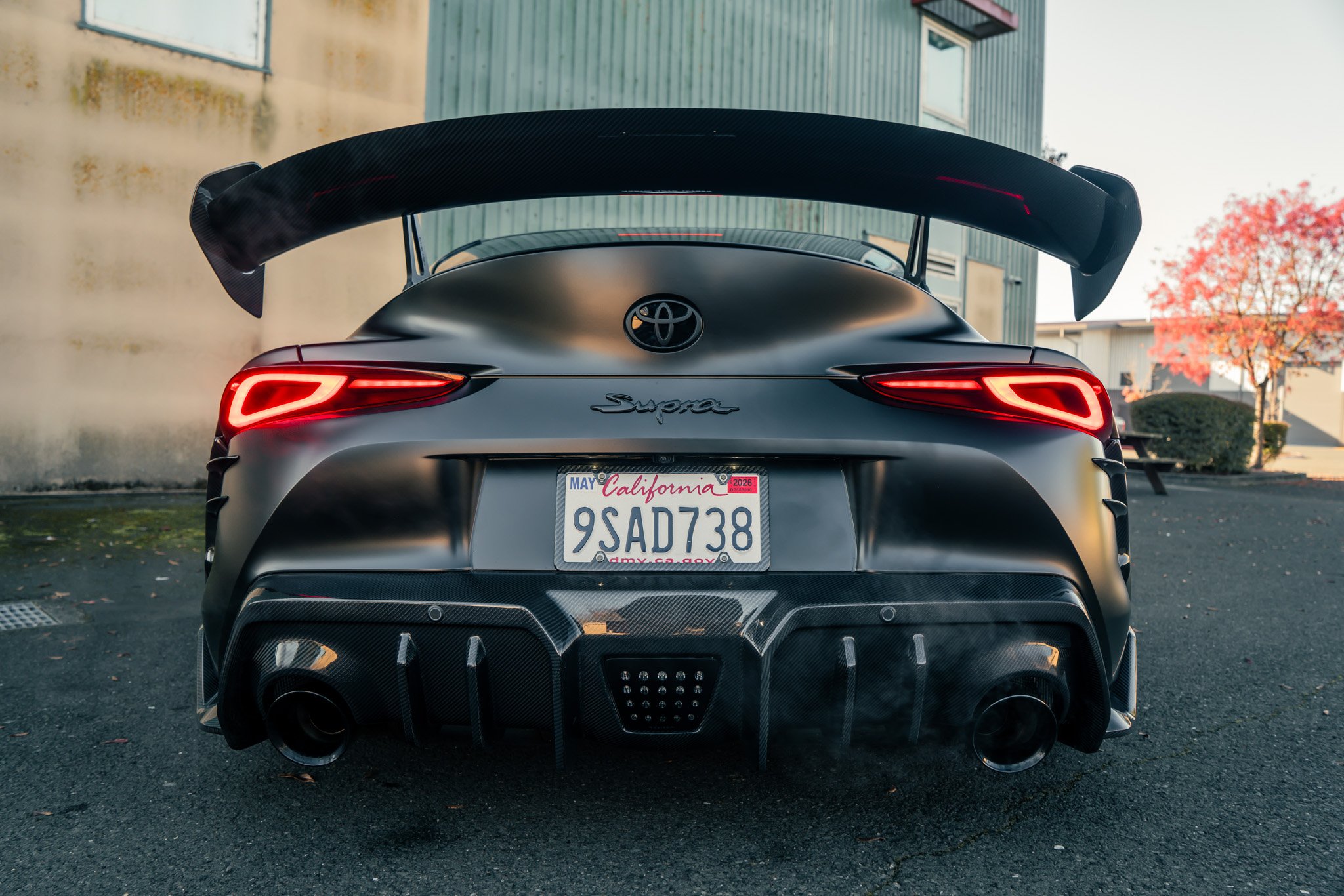 Rear view of a black Toyota Supra sports car with a large rear spoiler, carbon fiber diffuser, and red taillights, parked outdoors near industrial buildings with trees showing fall foliage.