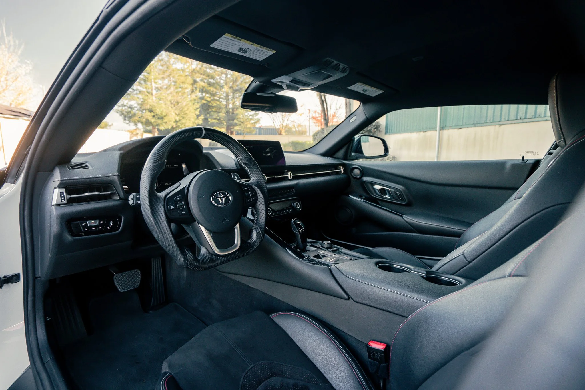 Interior of a modern car showing the steering wheel, dashboard, and front seats, with outside trees visible through the windows.