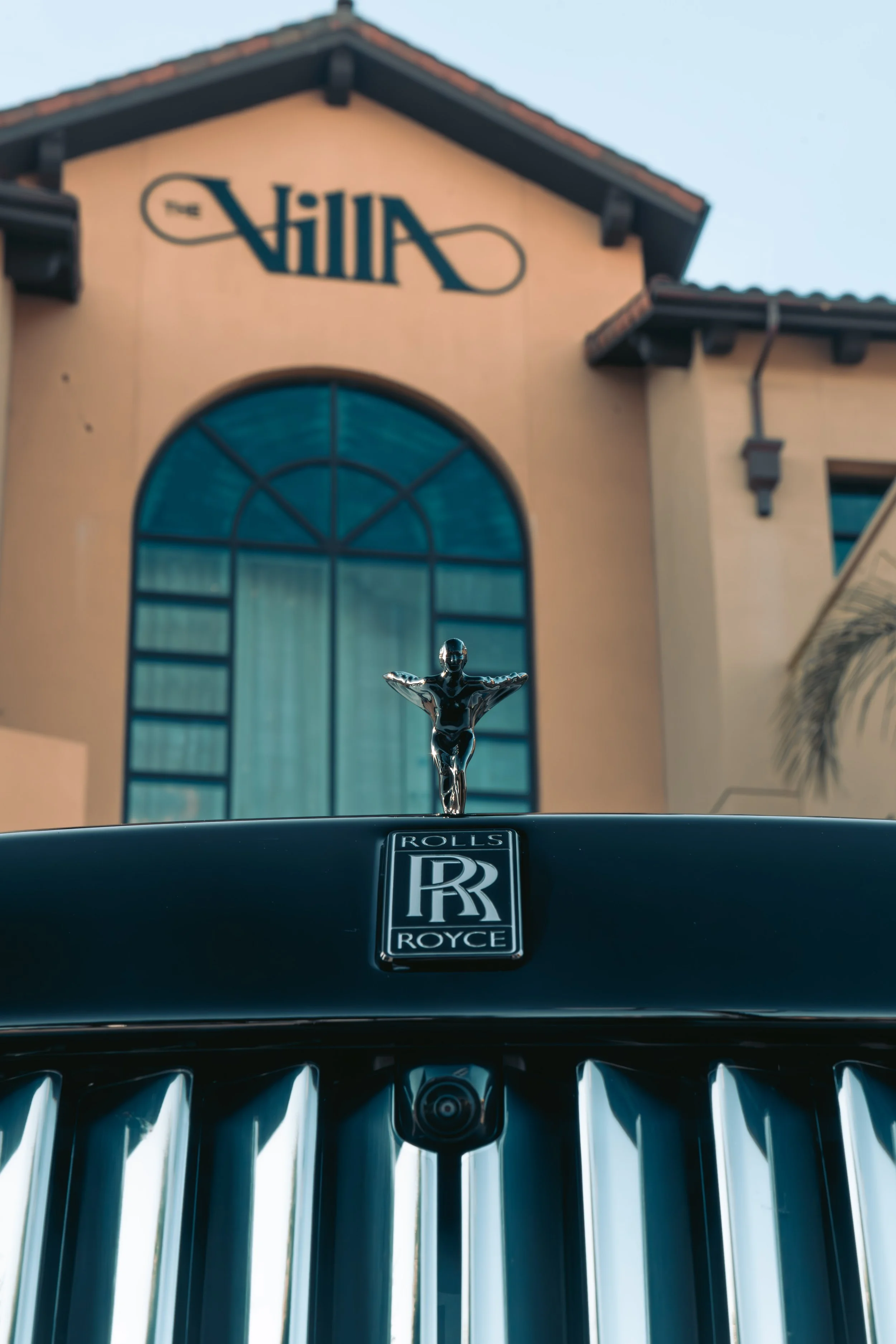 Close-up of the front of a Rolls Royce car, showing the iconic hood ornament and grille, with a building labeled 'The Villa' in the background.