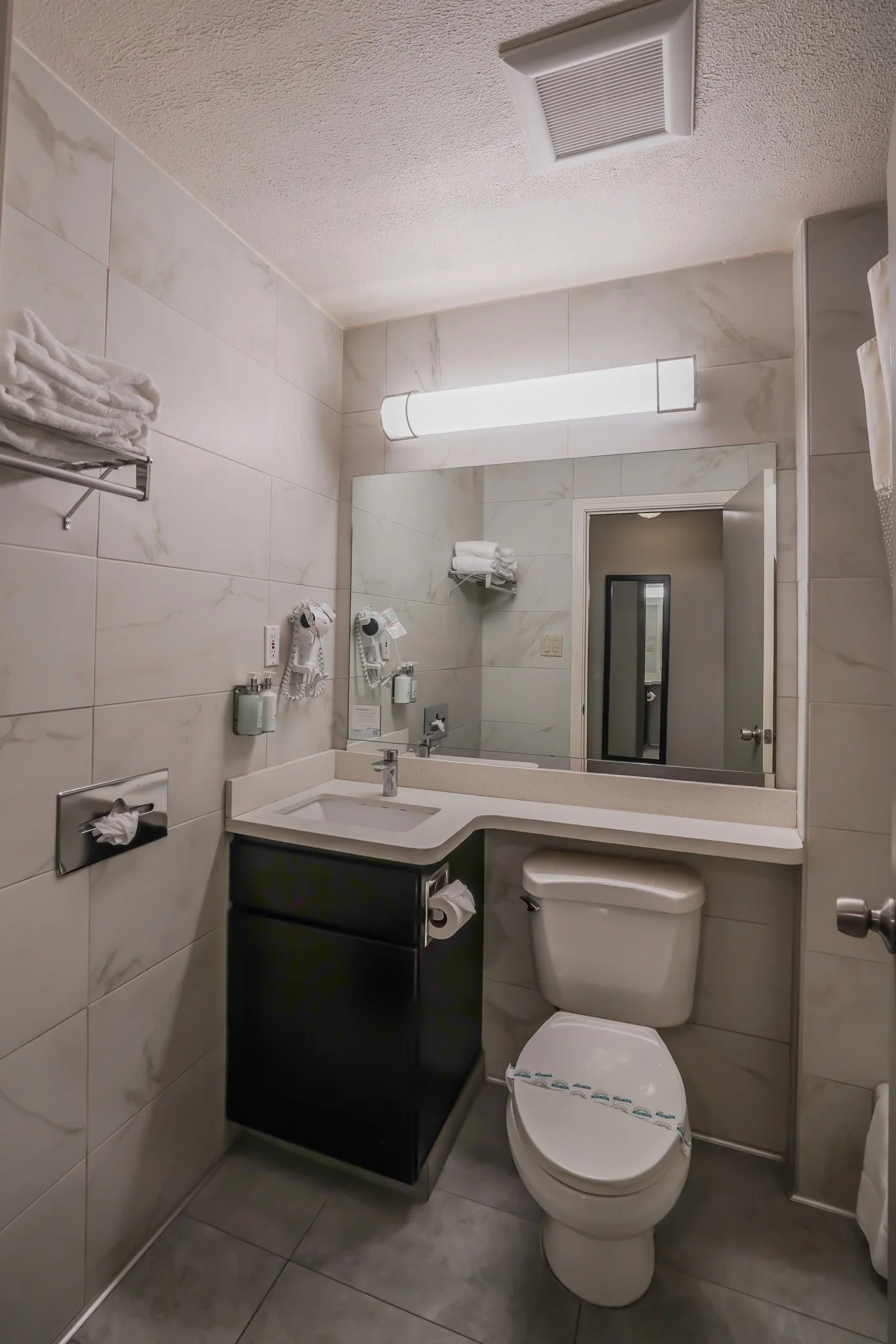 A small hotel bathroom with a marble tile wall, white rectangular mirror, black cabinet with a rolled toilet paper, white toilet, small white sink, mounted hairdryer, wall-mounted soap dispensers, towel shelf with towels, overhead light, and door ref