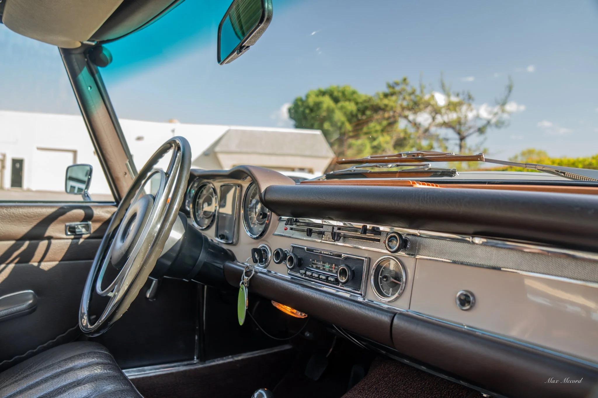 Interior of a vintage car showing the steering wheel, dashboard, and gauges, with a view outside through the windshield featuring trees and a clear sky.