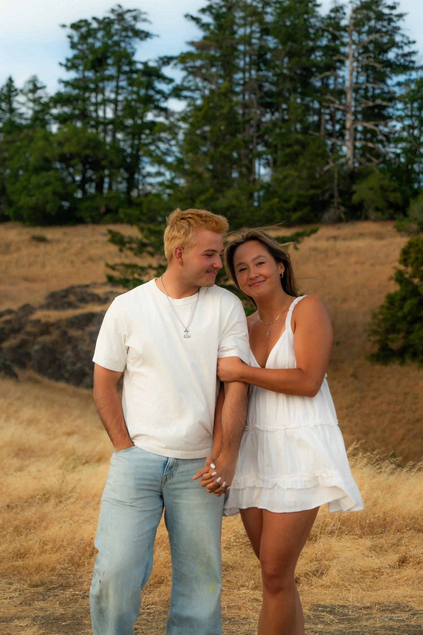 A young couple standing hand in hand outdoors on a grassy field with trees in the background, sharing a tender moment.