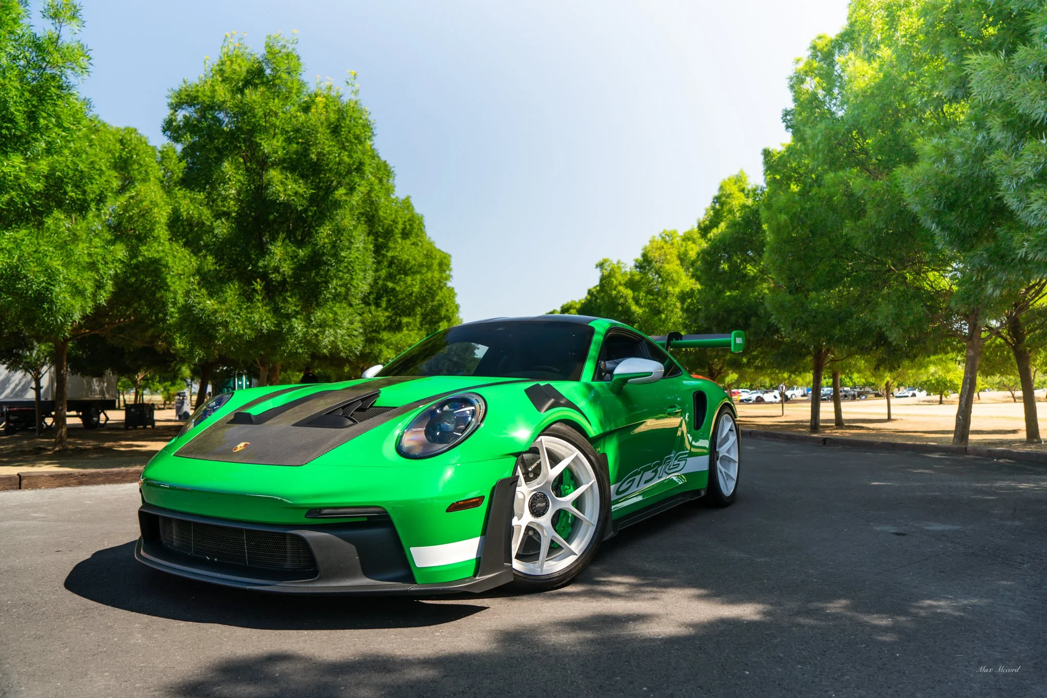 A bright green Porsche 911 GT3 racing car with black accents and white wheels parked on an asphalt surface under green trees on a sunny day.