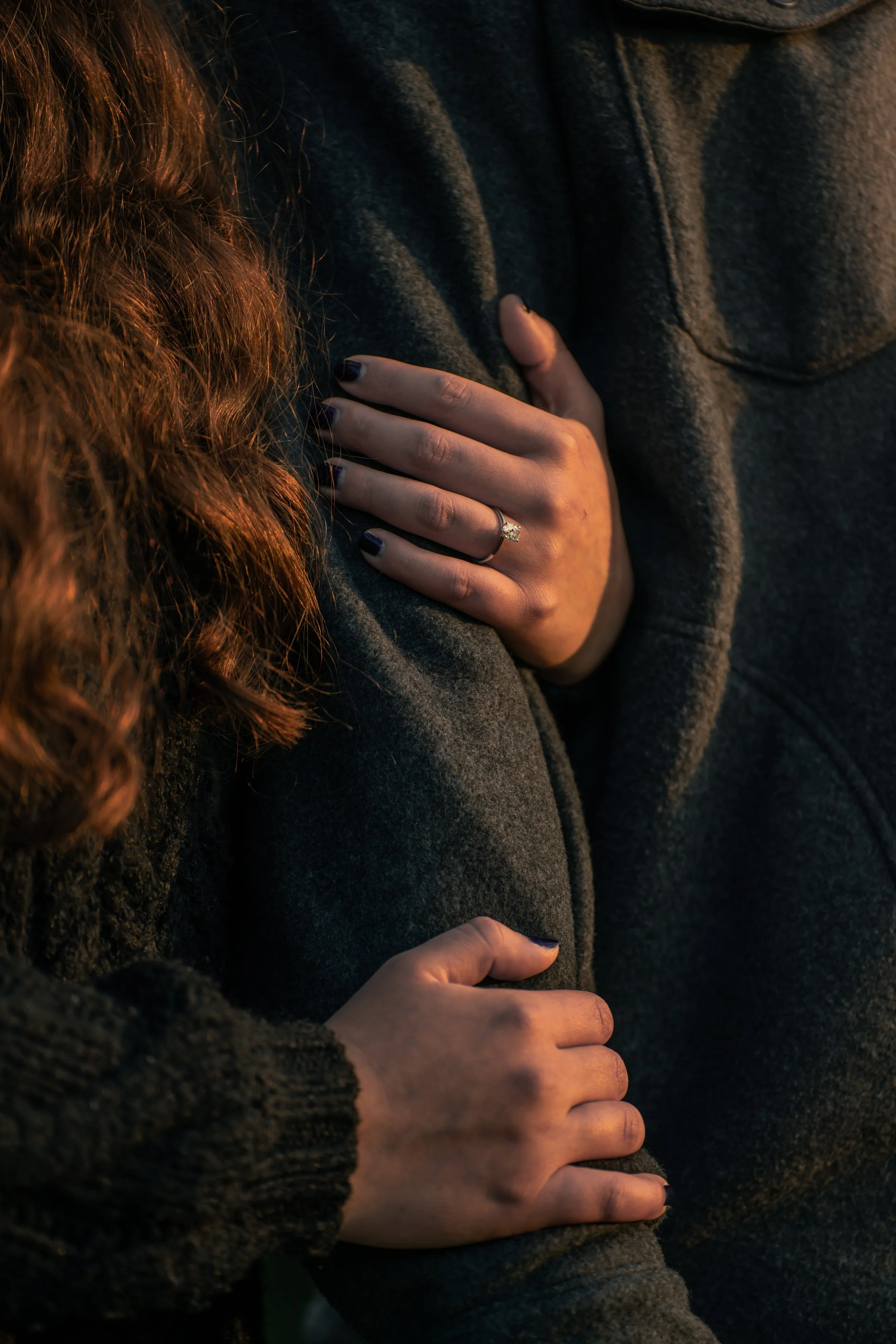 Close-up of a woman’s hand with a wedding ring resting on a man’s shoulder, both dressed in dark clothing.