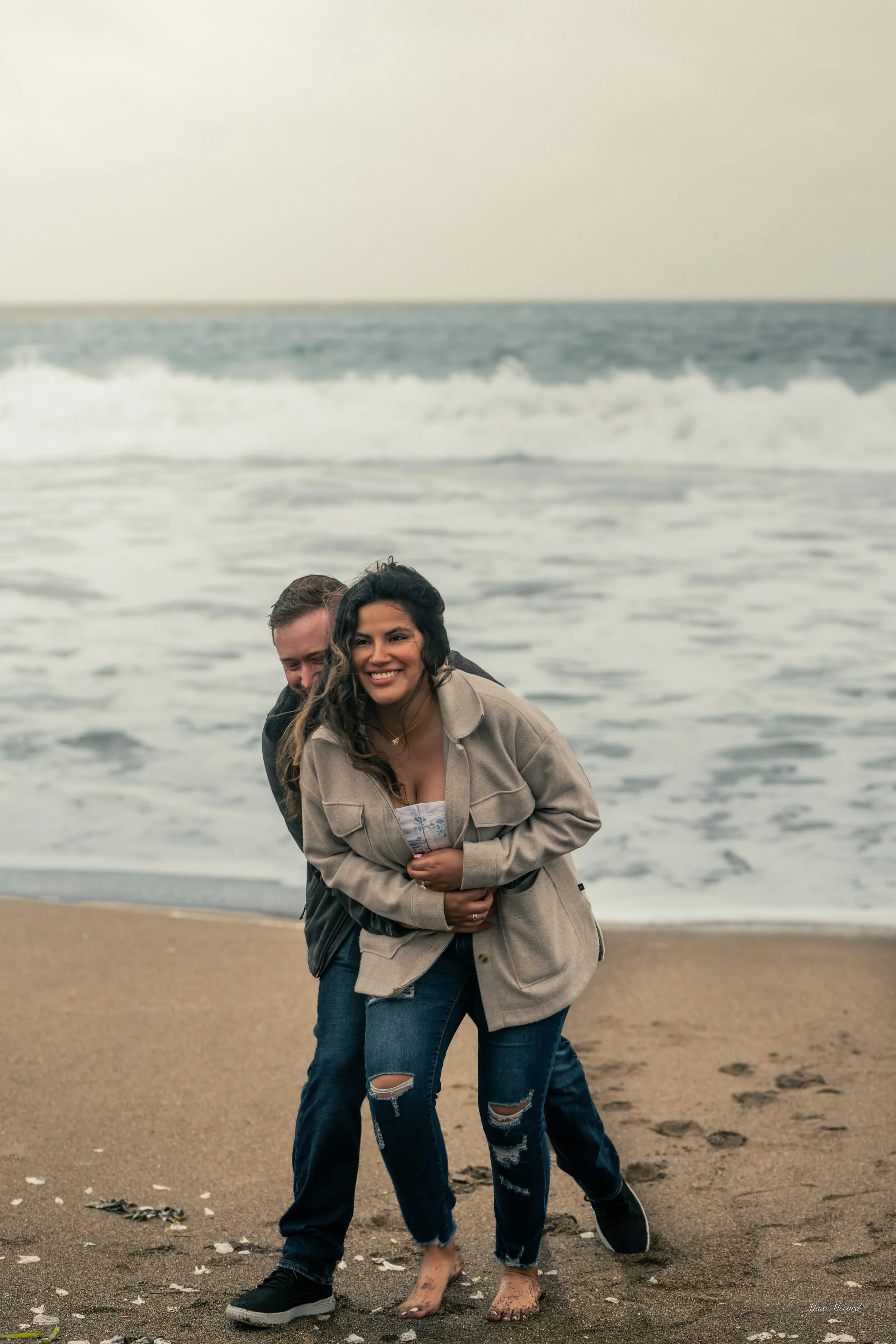 Two people, a man and a woman, enjoying time on a beach in front of the ocean. The woman is smiling, wearing a beige jacket and ripped jeans, standing barefoot in the sand. The man is partially behind her, wearing a dark jacket and jeans, hugging her