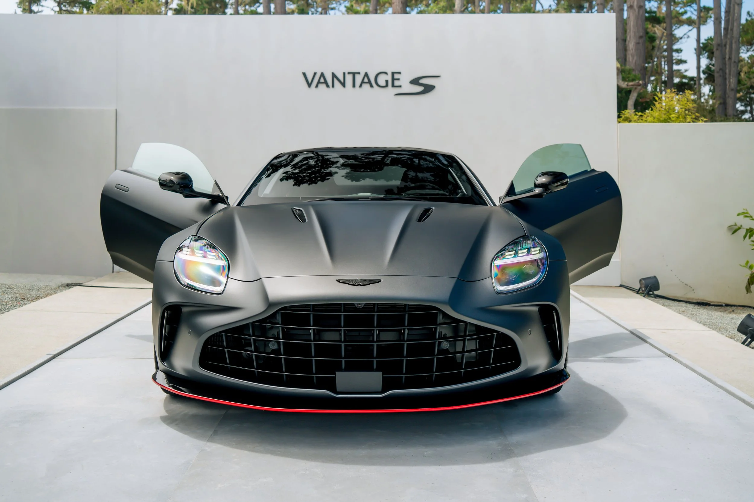 A matte black Aston Martin sports car with its doors open, parked on a display platform outdoors, with a white wall and trees in the background.