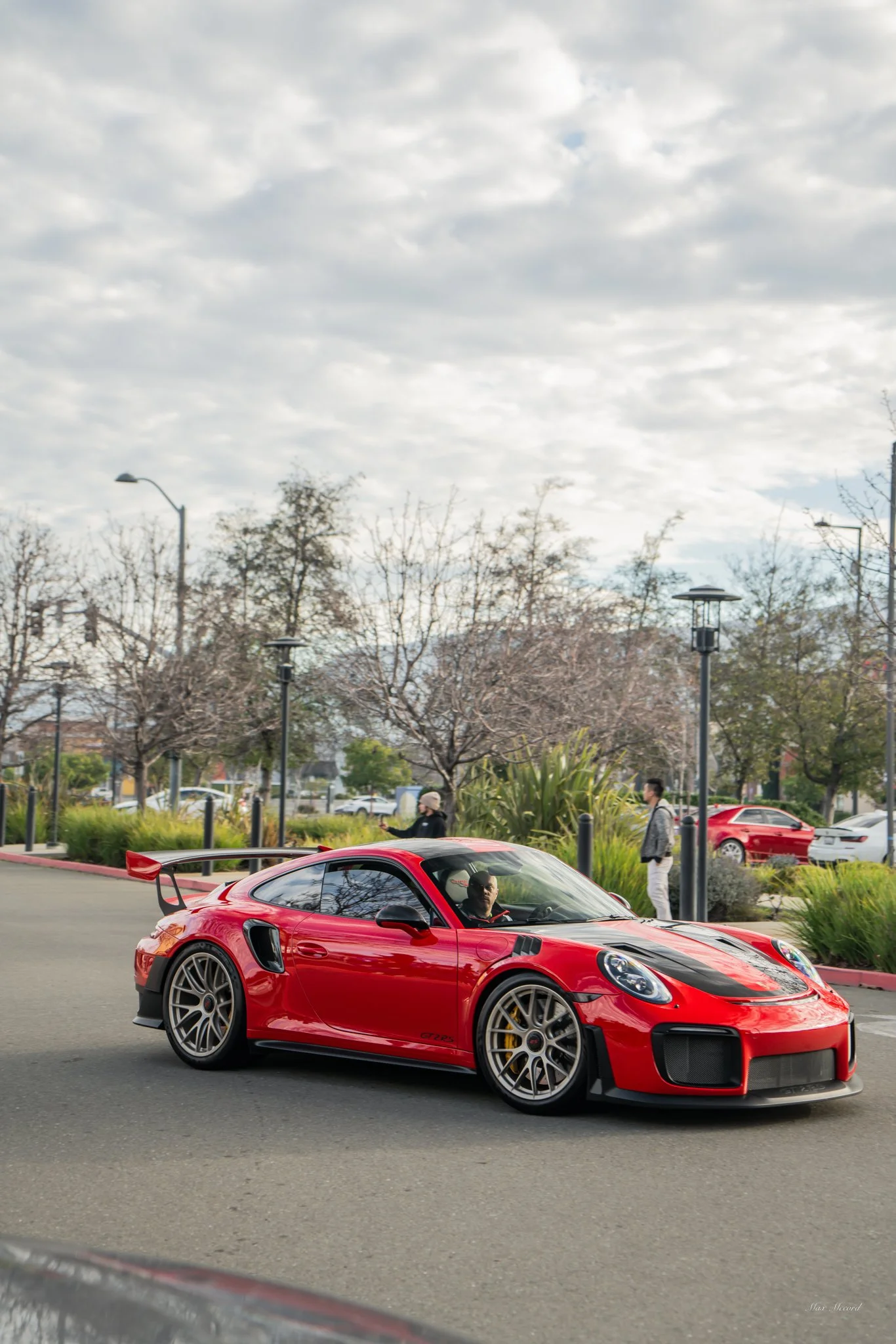 A red sports car with black racing stripes and a rear spoiler parked on a street, with trees and pedestrians in the background.