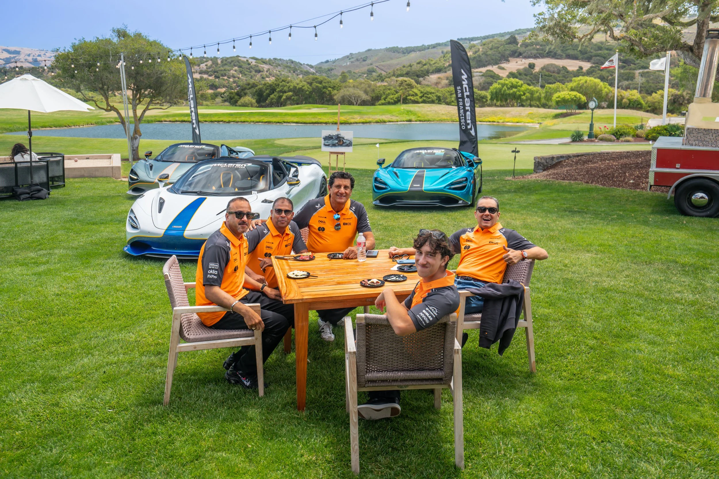 Group of five men wearing orange and black racing team shirts sitting at a wooden table outdoors, with two high-performance sports cars in the background on a green lawn at a golf course, near a lake with hills and trees.