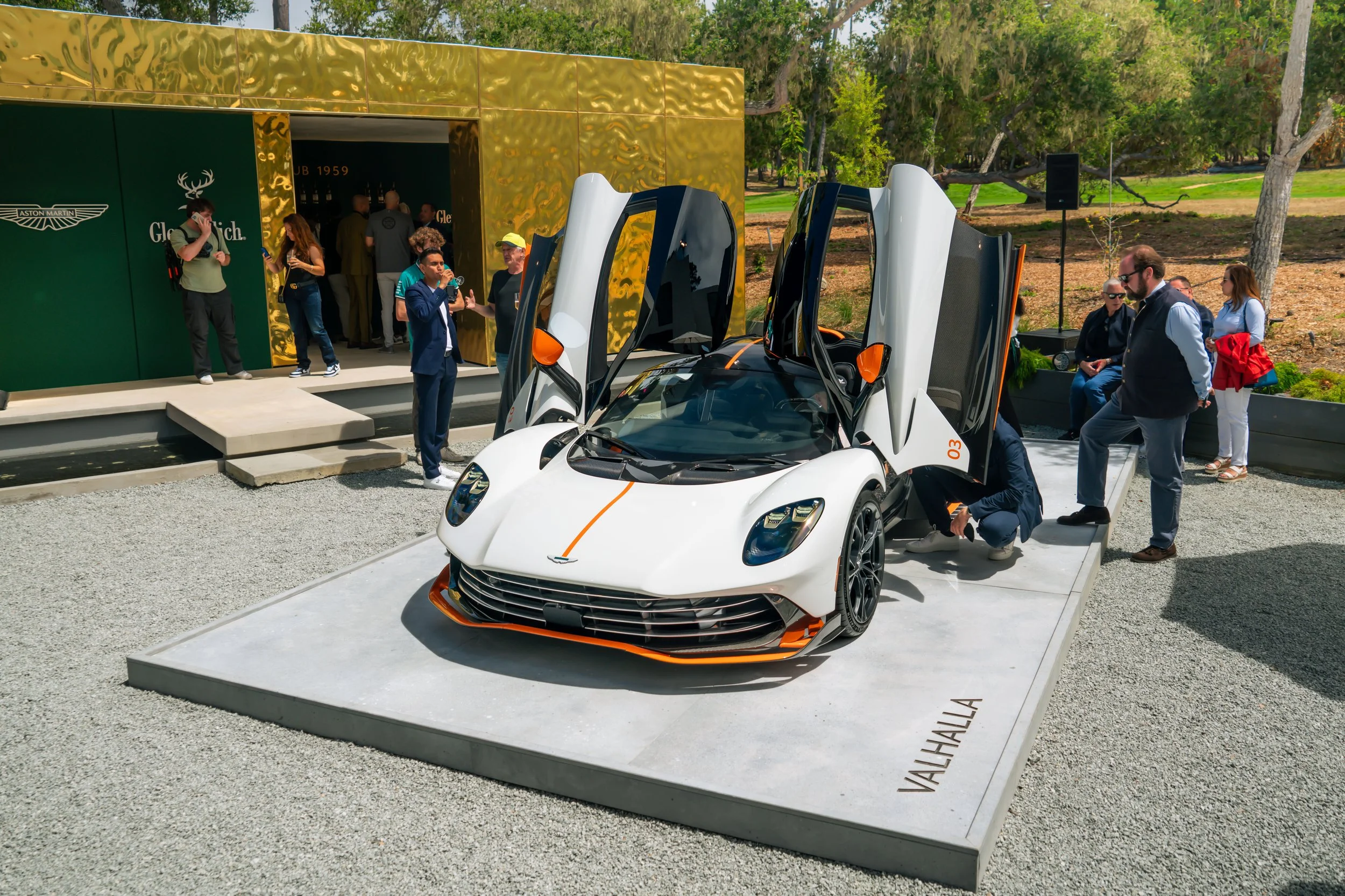 A white Valhalla sports car with open doors displayed on a platform outdoors, surrounded by people at an event, with a green and gold building and trees in the background.
