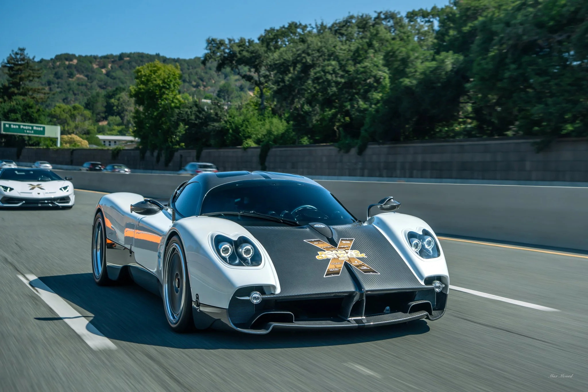 A silver and black high-performance sports car with a large X logo on the front, driving on a highway with other cars, surrounded by green trees and hill scenery in the background.