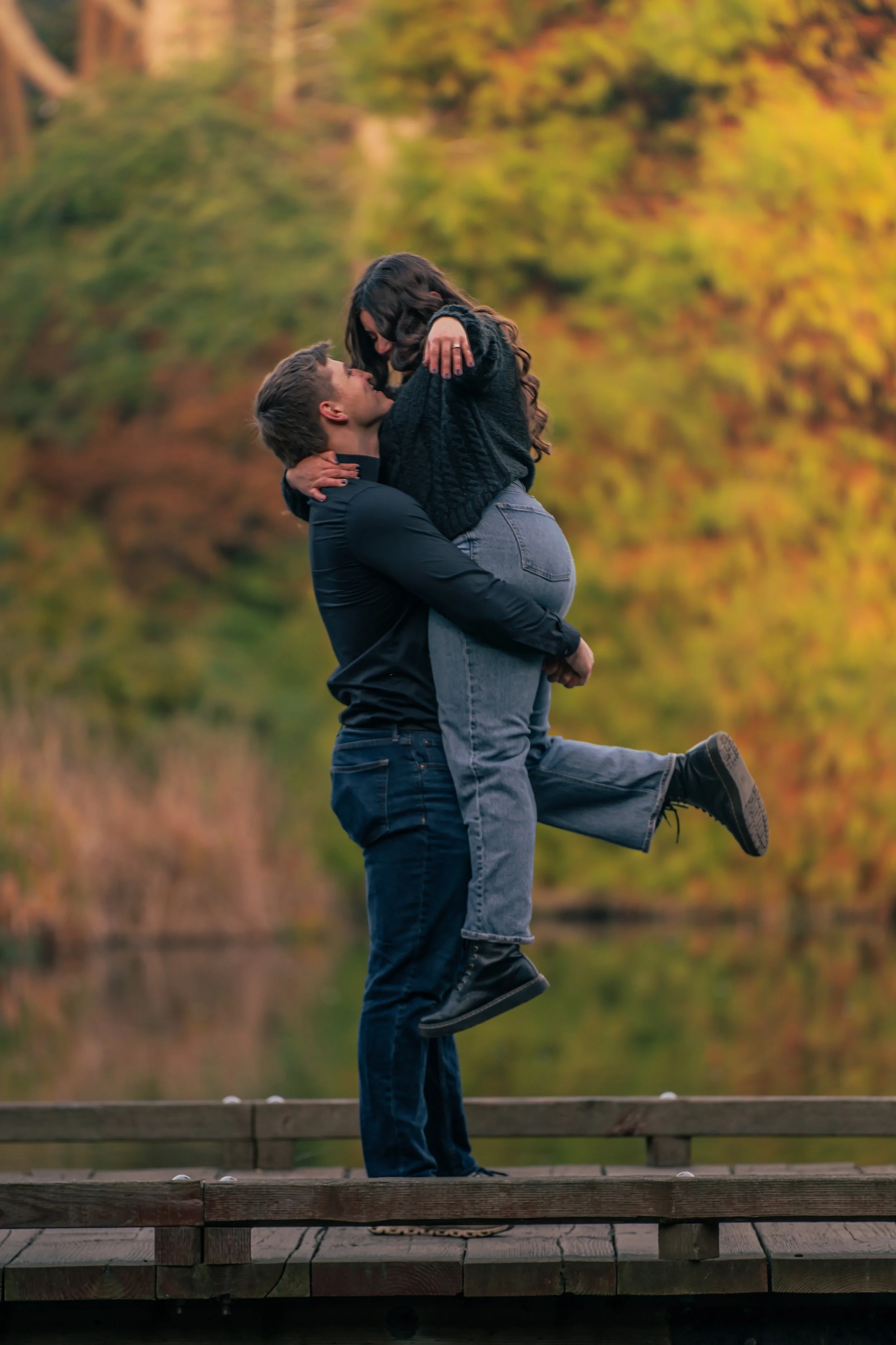 A couple on a wooden dock, with the man lifting the woman in his arms as they share a kiss, surrounded by fall foliage.