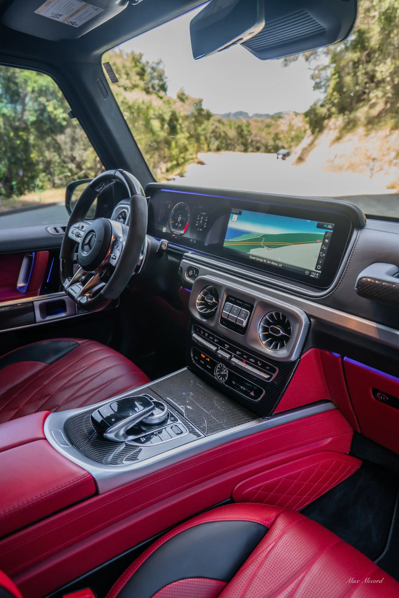 Interior of a luxury car with red leather seats, a digital dashboard, and a navigation system displaying a map, driving along a scenic road with greenery and hills.