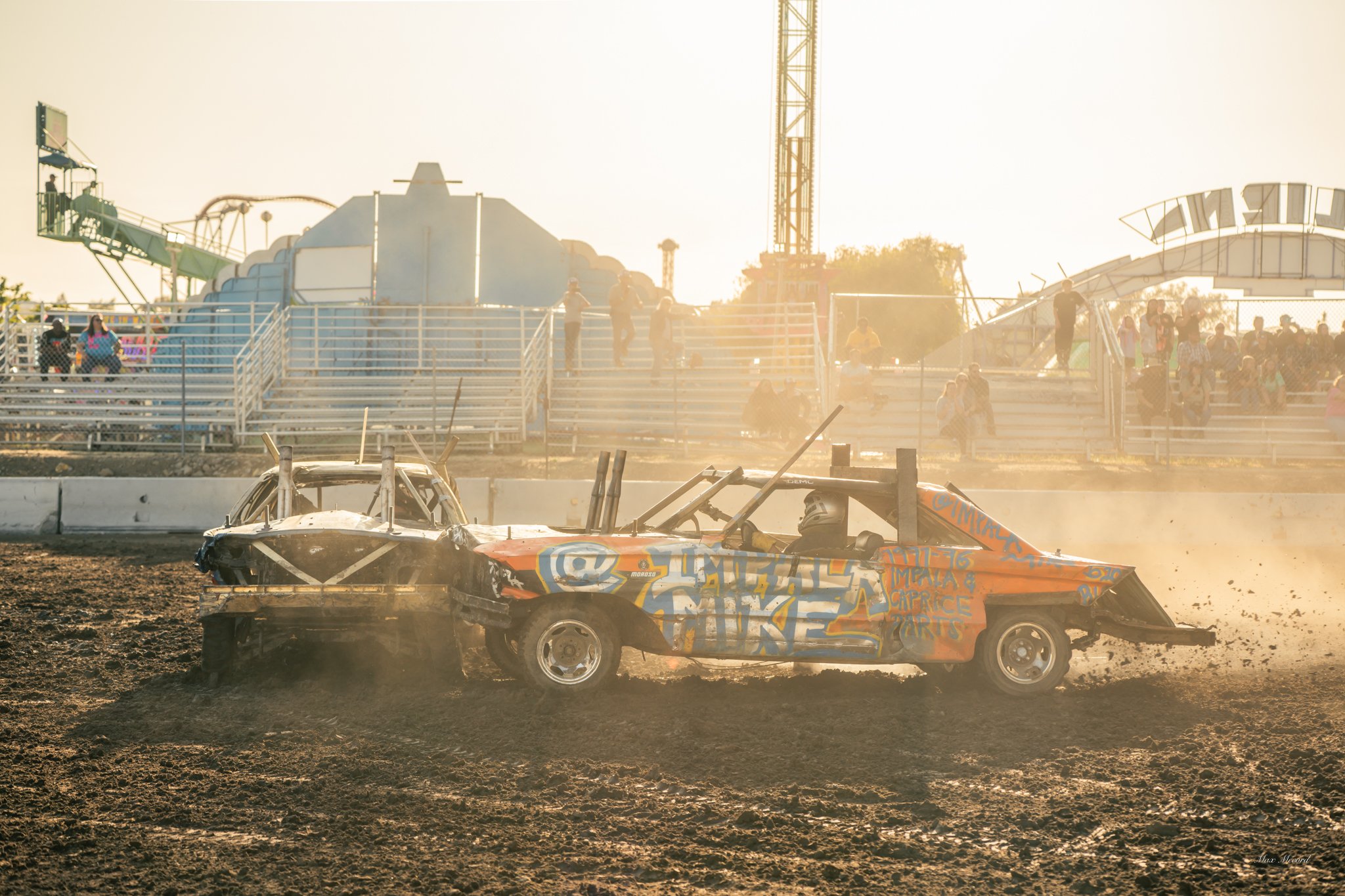 A race car and a wrecked car on a dirt track during a demolition derby, with spectators watching from bleachers in the background.
